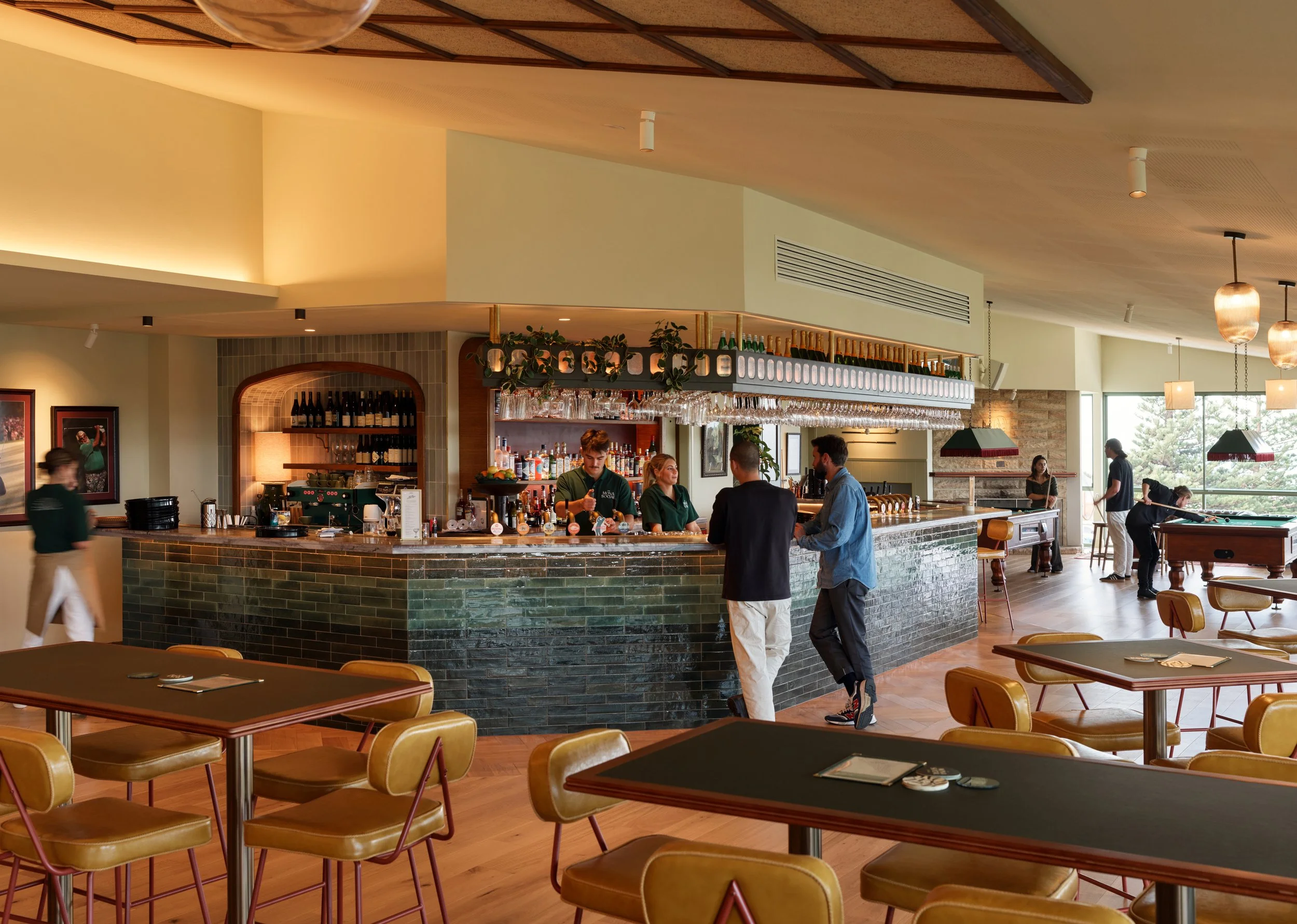 Interior of a modern bar and lounge area featuring a curved bar counter with stools, a few people sitting and talking, and a pool table in the background. The space is well-lit with natural light from large windows, and decorated with hanging pendant lights and framed pictures on the walls.