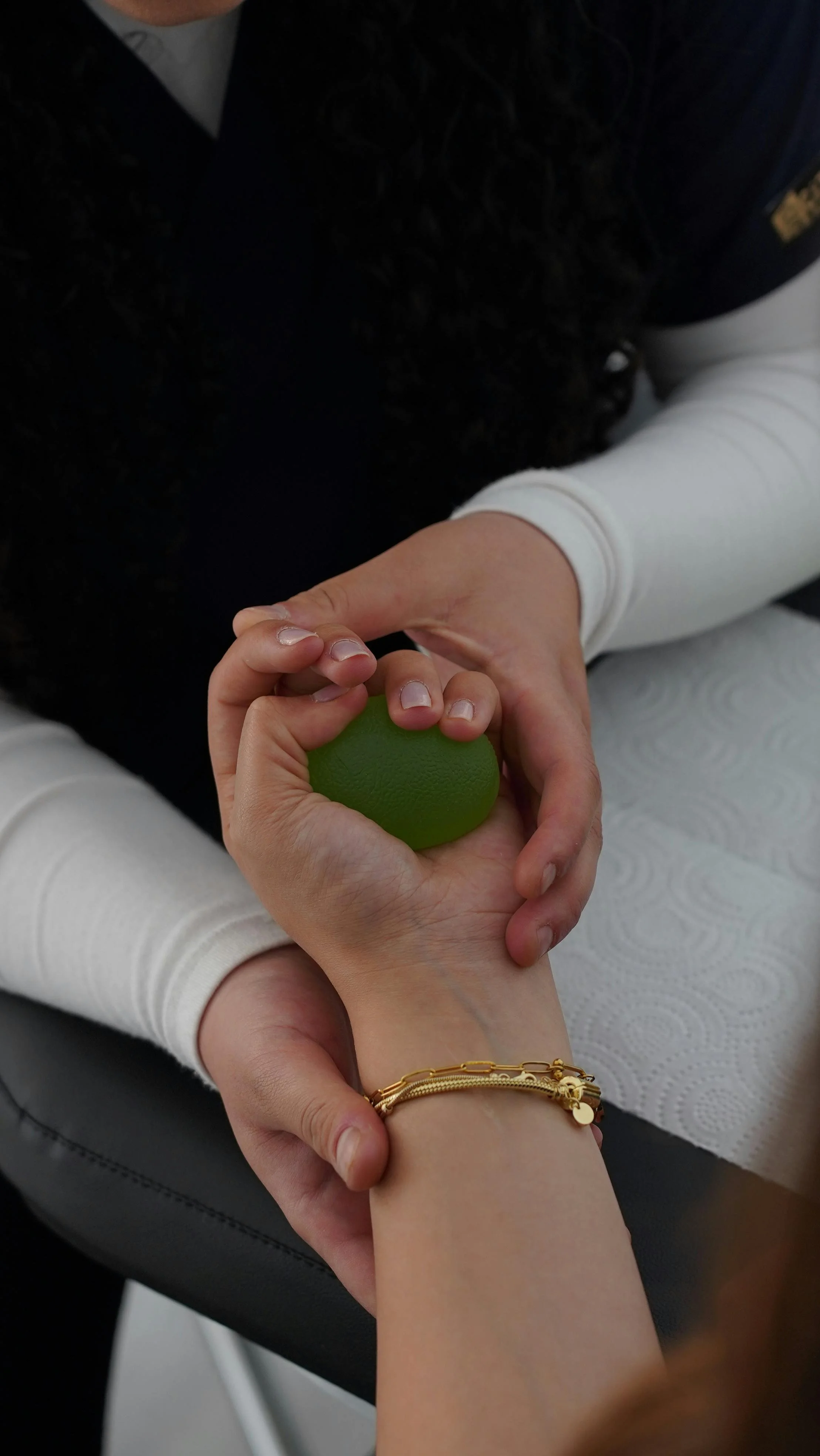 Person receiving a hand massage with a green stress ball, wearing a gold bracelet, sitting on a white paper-covered surface.