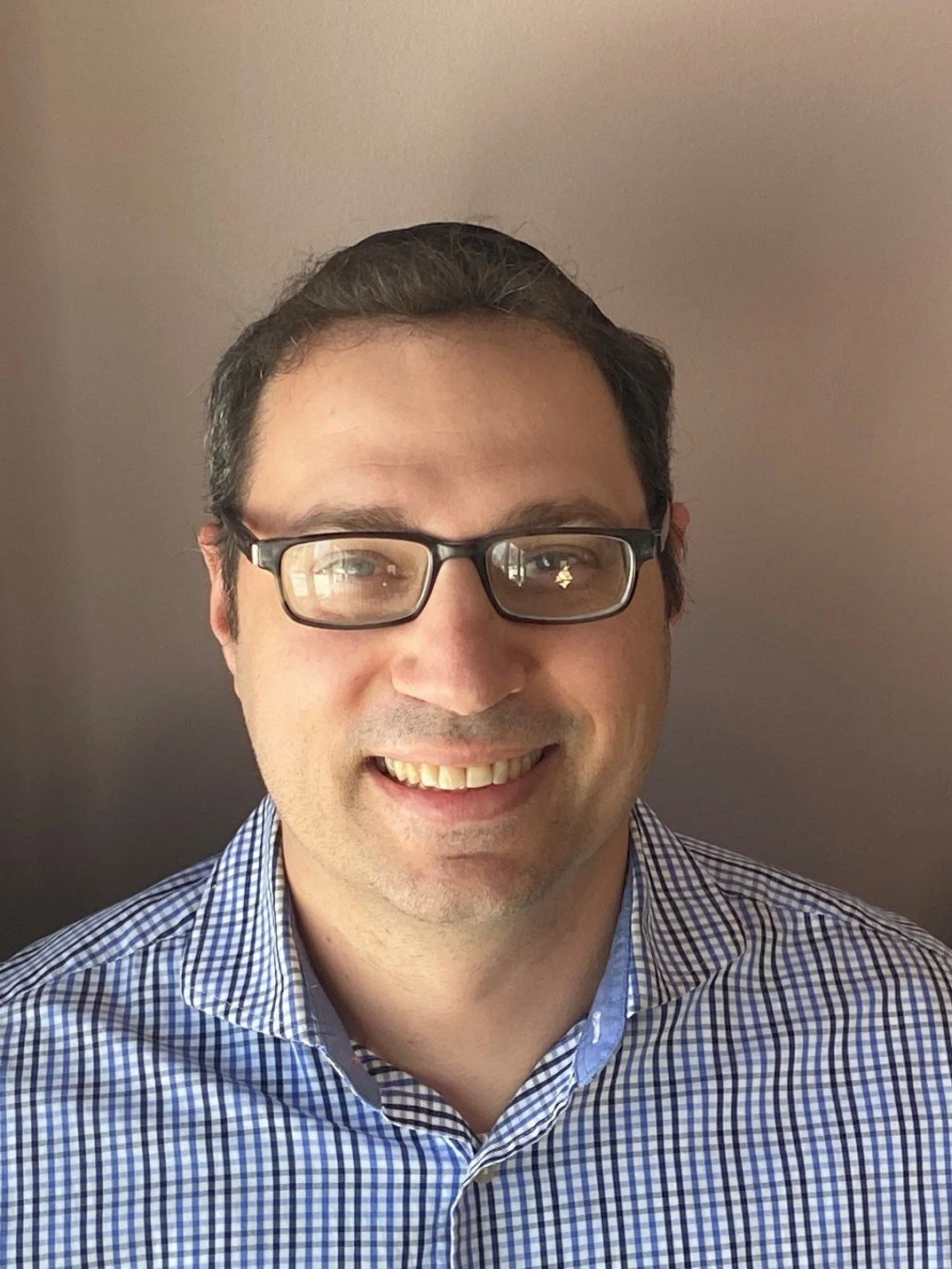 A man with dark hair, glasses, and a checkered shirt smiling, standing in front of a plain wall.