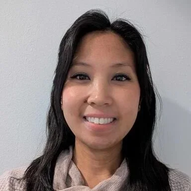 A woman with long dark hair smiling at the camera against a plain gray background.