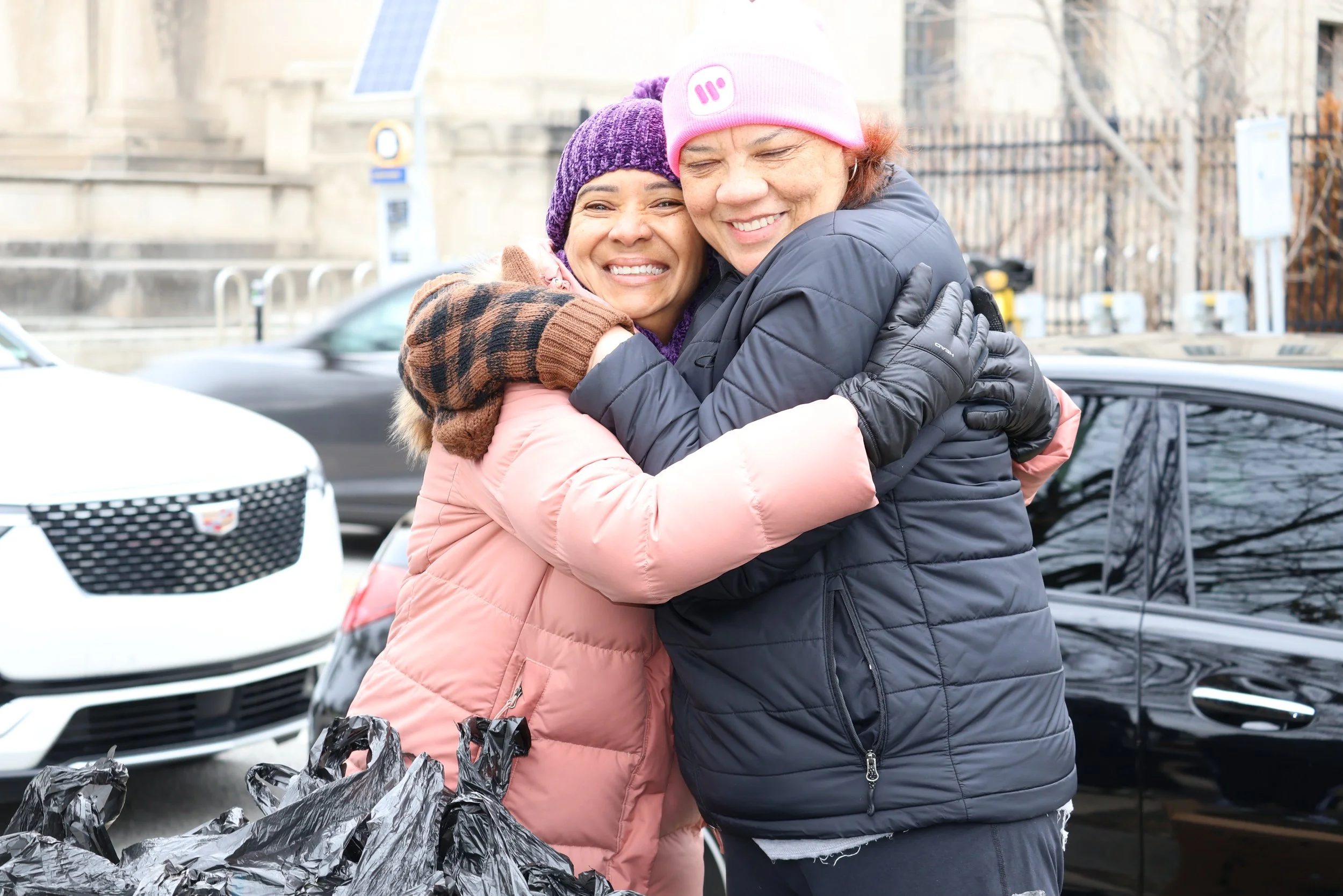 Two women hugging each other outdoors, smiling happily. One woman is wearing a pink jacket and purple knit hat, and the other is wearing a black jacket and pink knit hat. There are cars and a building in the background.