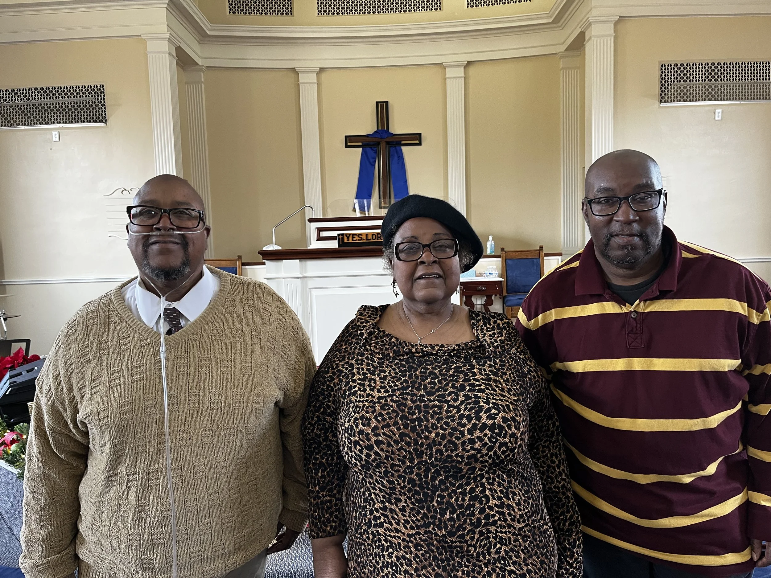Three adults stand in front of an altar with a cross draped in a blue cloth inside a church. The man on the left wears glasses, a beige sweater, and a white shirt. The woman in the middle wears glasses, a leopard print top, and a black hat. The man on the right wears glasses and a maroon and yellow striped polo shirt.