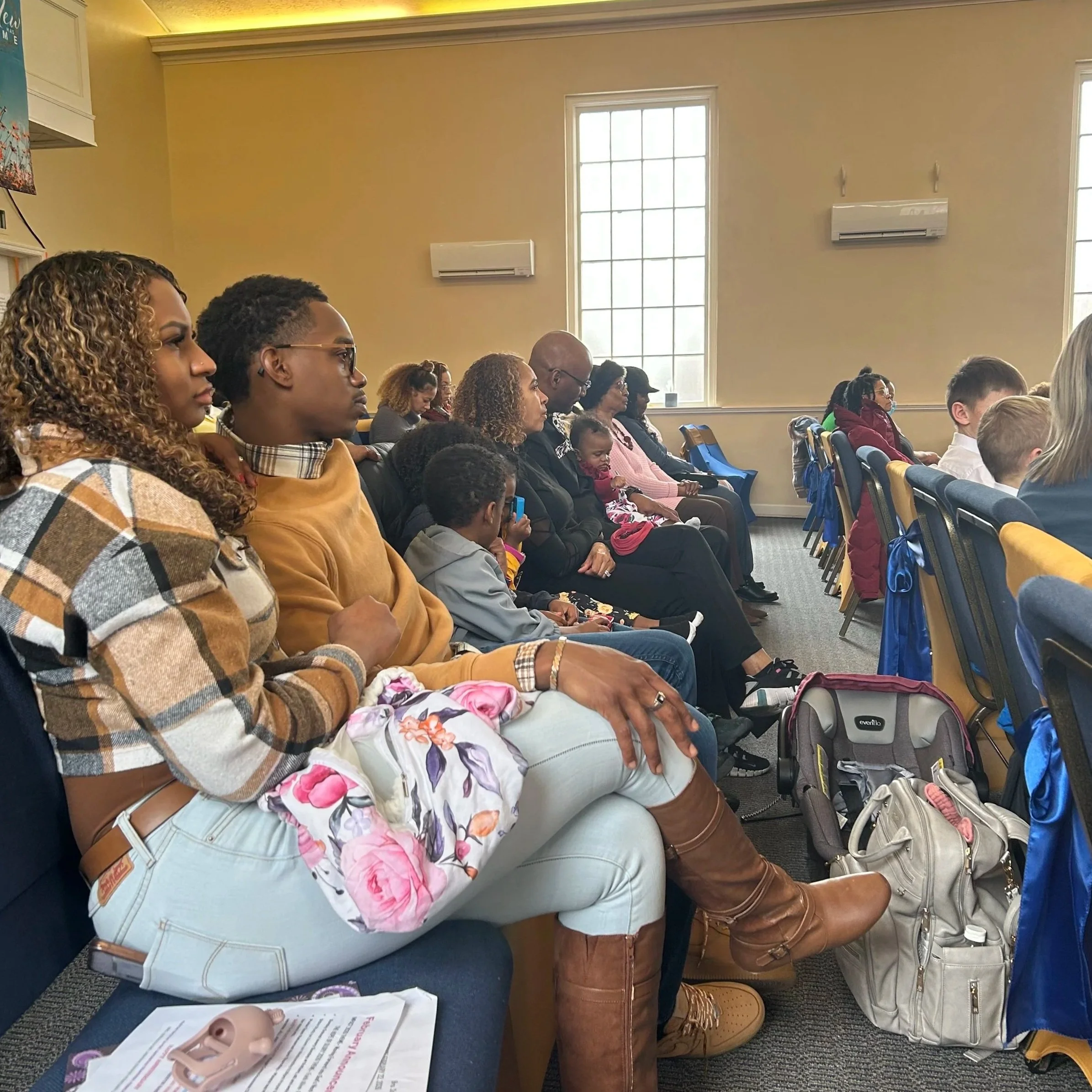 A diverse group of people sitting in a room attending a seminar or presentation, with some taking notes, and a window in the background.