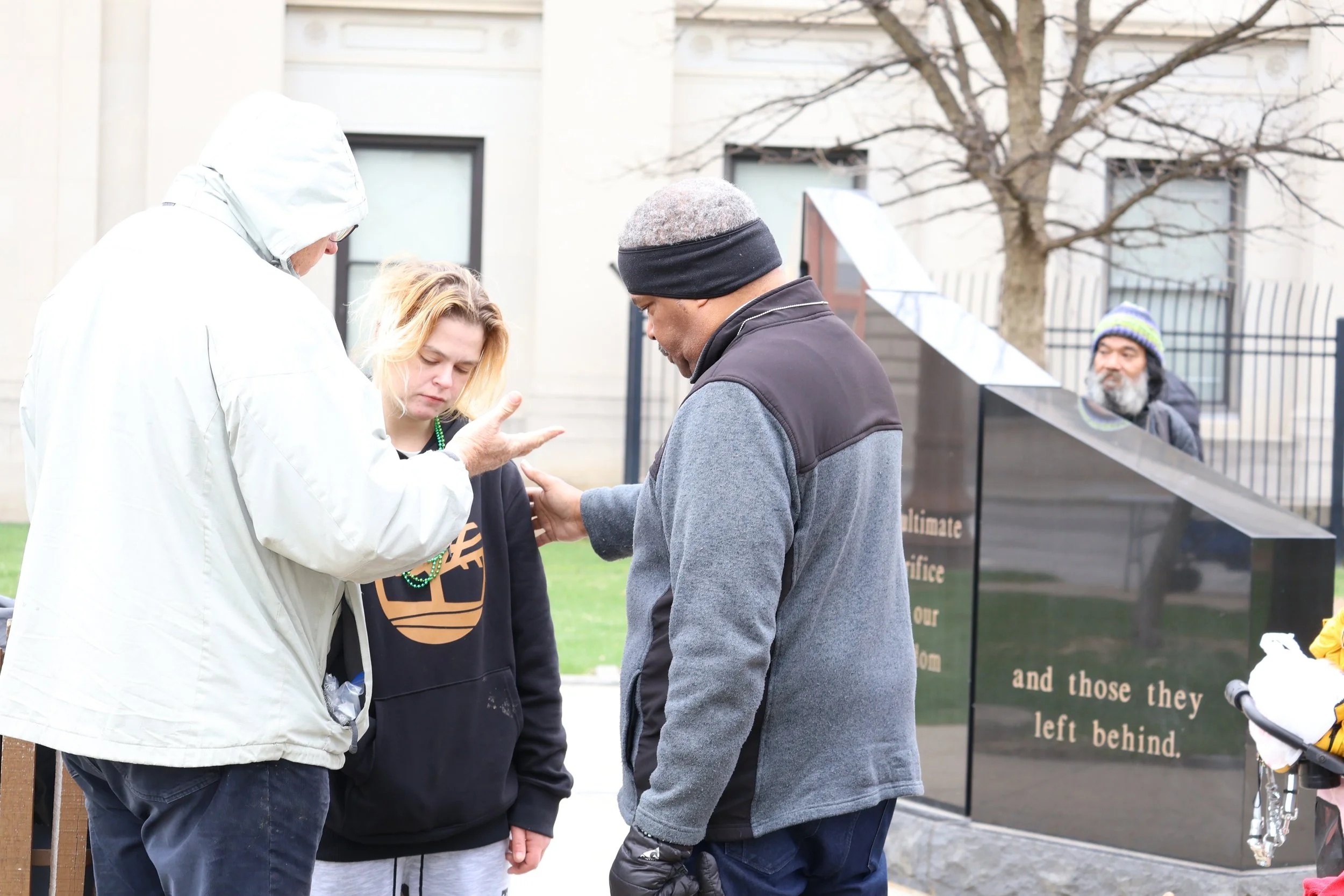 Three people, two men and one woman, appear to be engaged in a serious conversation outdoors near a large informational sign and a leafless tree, with a building in the background.