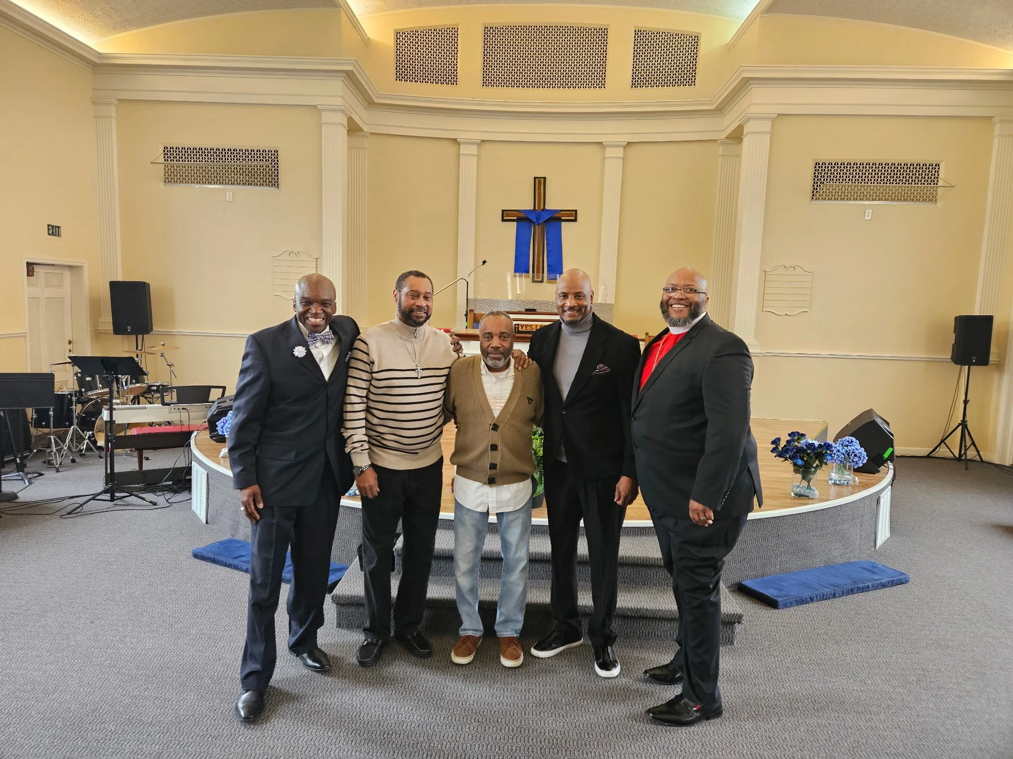 Five men standing together in a church, smiling for the camera. The church interior features a cross with blue cloth on the wall behind them, musical instruments on the left, and flower arrangements on the right. The men are dressed in formal and semi-formal attire, with one wearing a clerical collar.