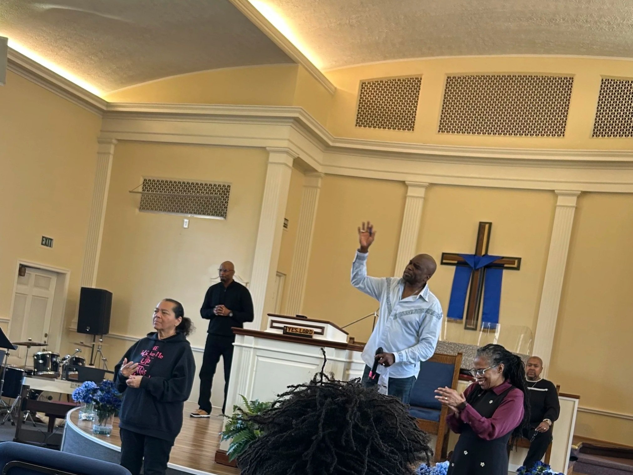 People gathered in a church during a service or event, some are standing and others are listening, with a man in a white shirt raising his hand, and a woman in black with a hoodie in the foreground.