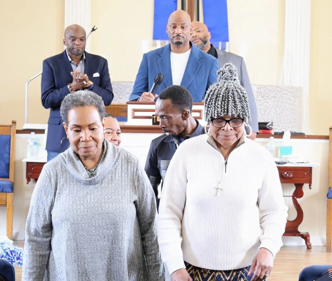 A group of people praying in a church, with two women in the front and three men behind them, one of whom is holding a microphone.