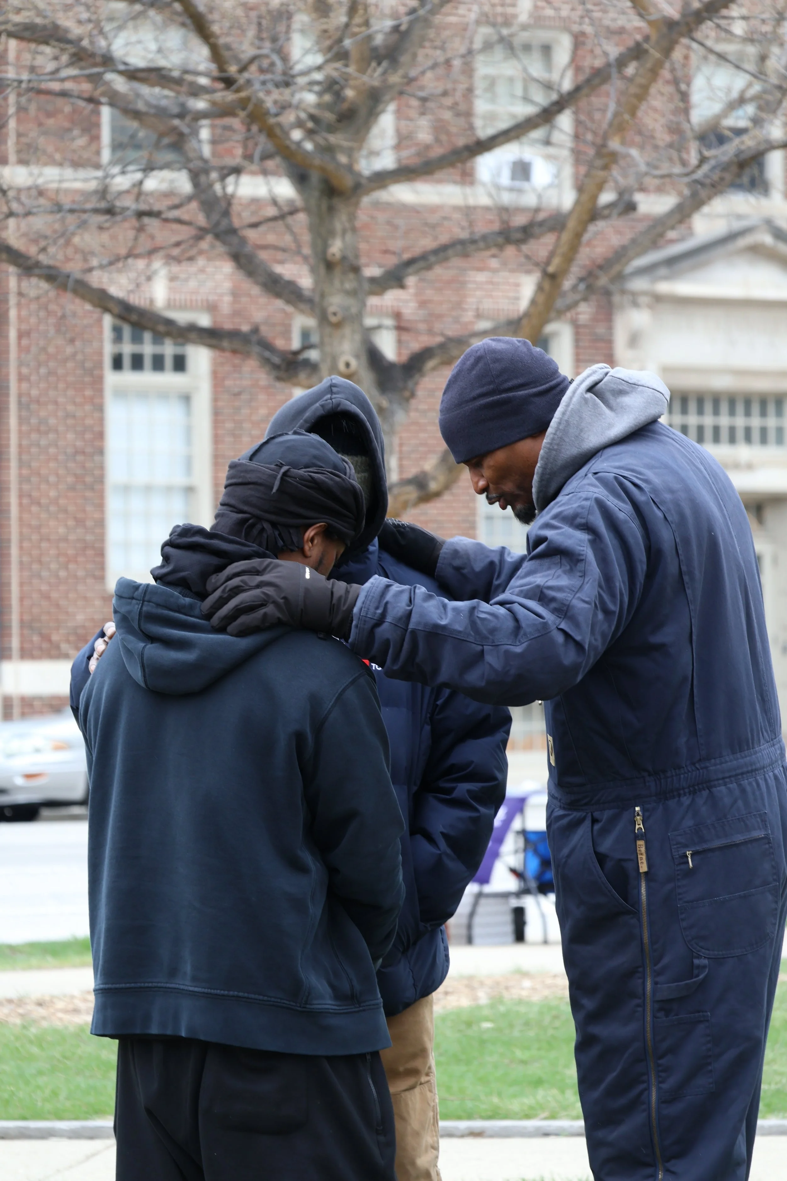 Three men standing closely together outdoors with their heads bowed, appearing to be praying or in reflection, with a bare tree and brick building in the background.