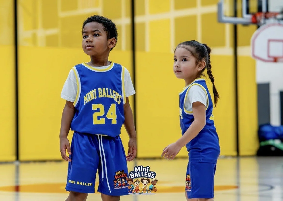Two young children in blue basketball uniforms on a gymnasium court, with a yellow wall and basketball hoop in the background, participating in a basketball game or practice.