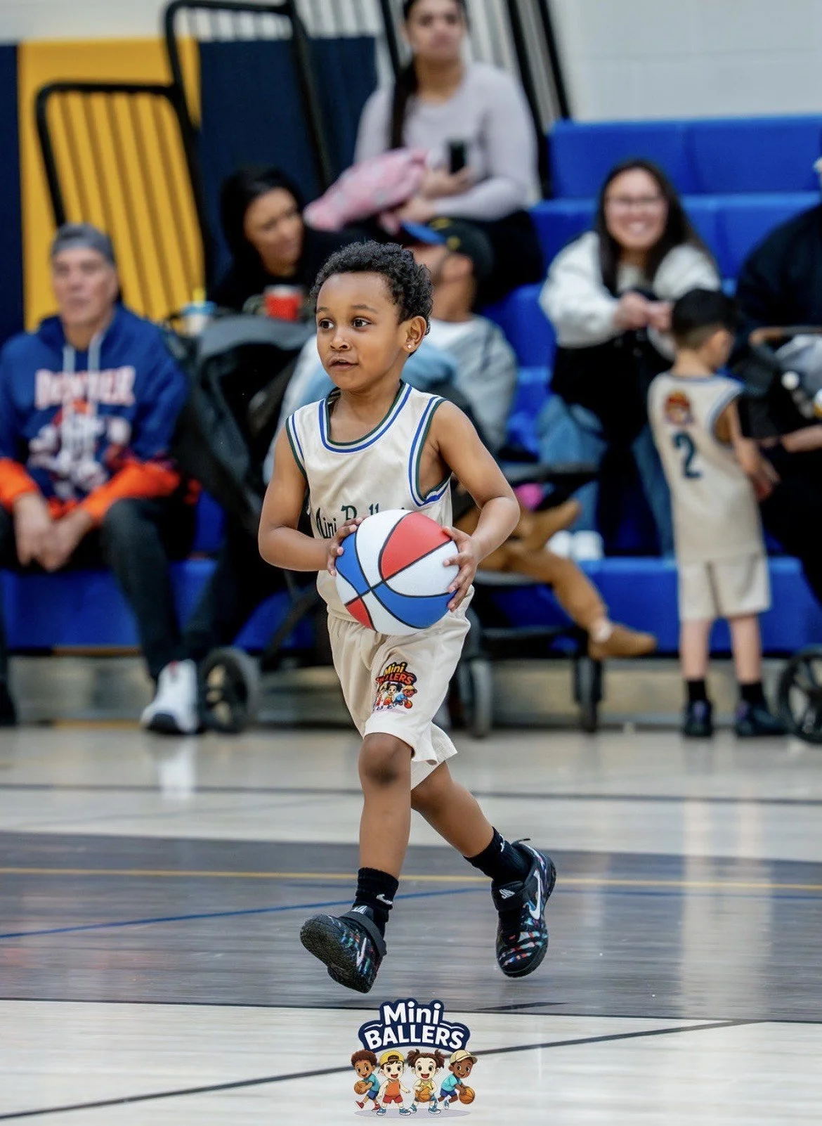 A young boy playing basketball in a gymnasium, holding a colorful basketball, with spectators seated in the background.