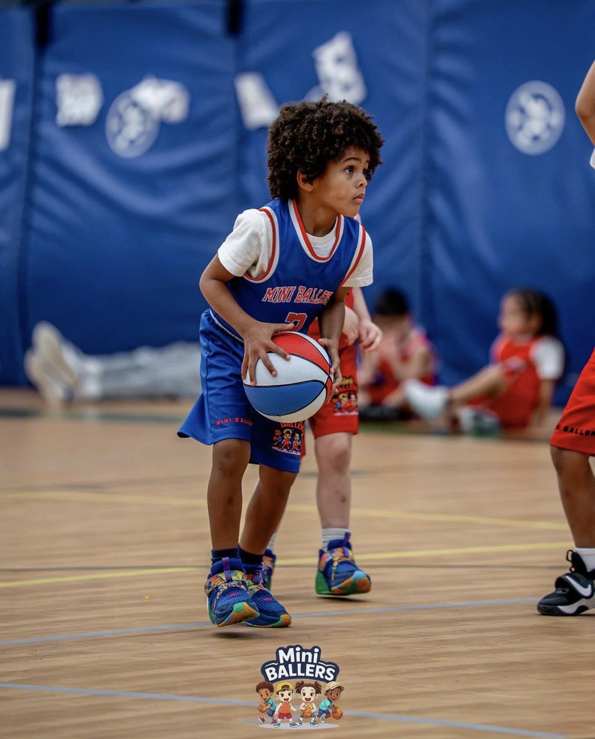 Young boy with curly hair holding a basketball on an indoor basketball court, wearing a blue and white sports uniform and colorful sneakers, with other children sitting in the background.