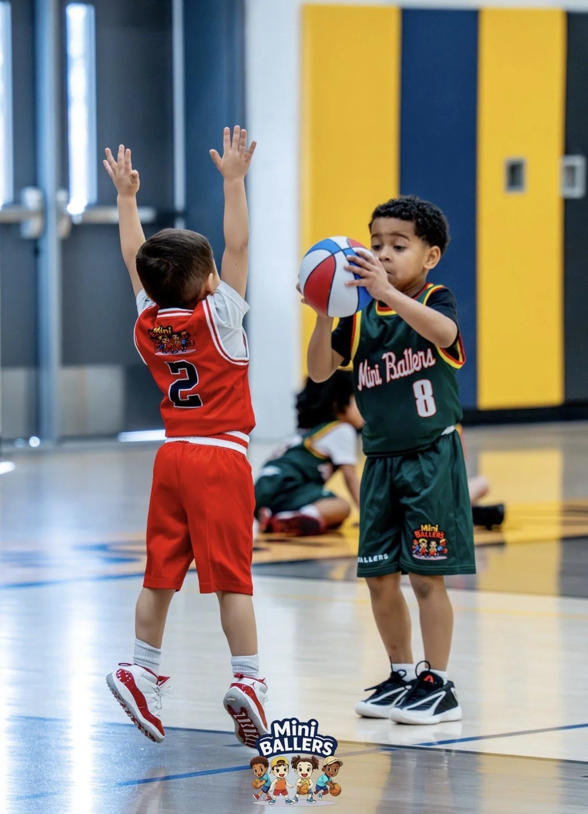 Two young boys playing basketball indoors, one with a red jersey and the other with a green jersey, with a logo that says 'Mini Ballers' on their jerseys. The boy in green is holding a colorful basketball, while the boy in red is blocking with his ar