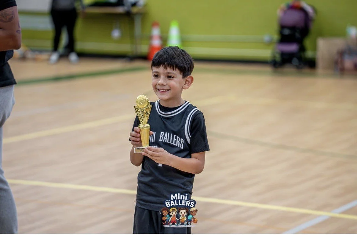 A young boy in a black sports jersey smiling and holding a gold trophy on an indoor basketball court.