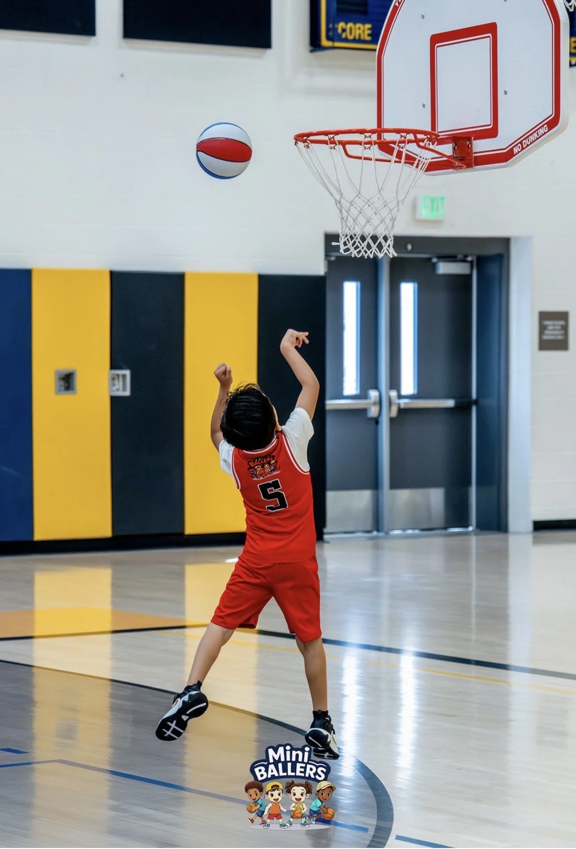 A young boy in a red and white basketball uniform with the number 5 is shooting a basketball in an indoor gymnasium. The ball is mid-air near the hoop, and the boy is facing away from the camera, standing on the court floor. The gym has a yellow and 