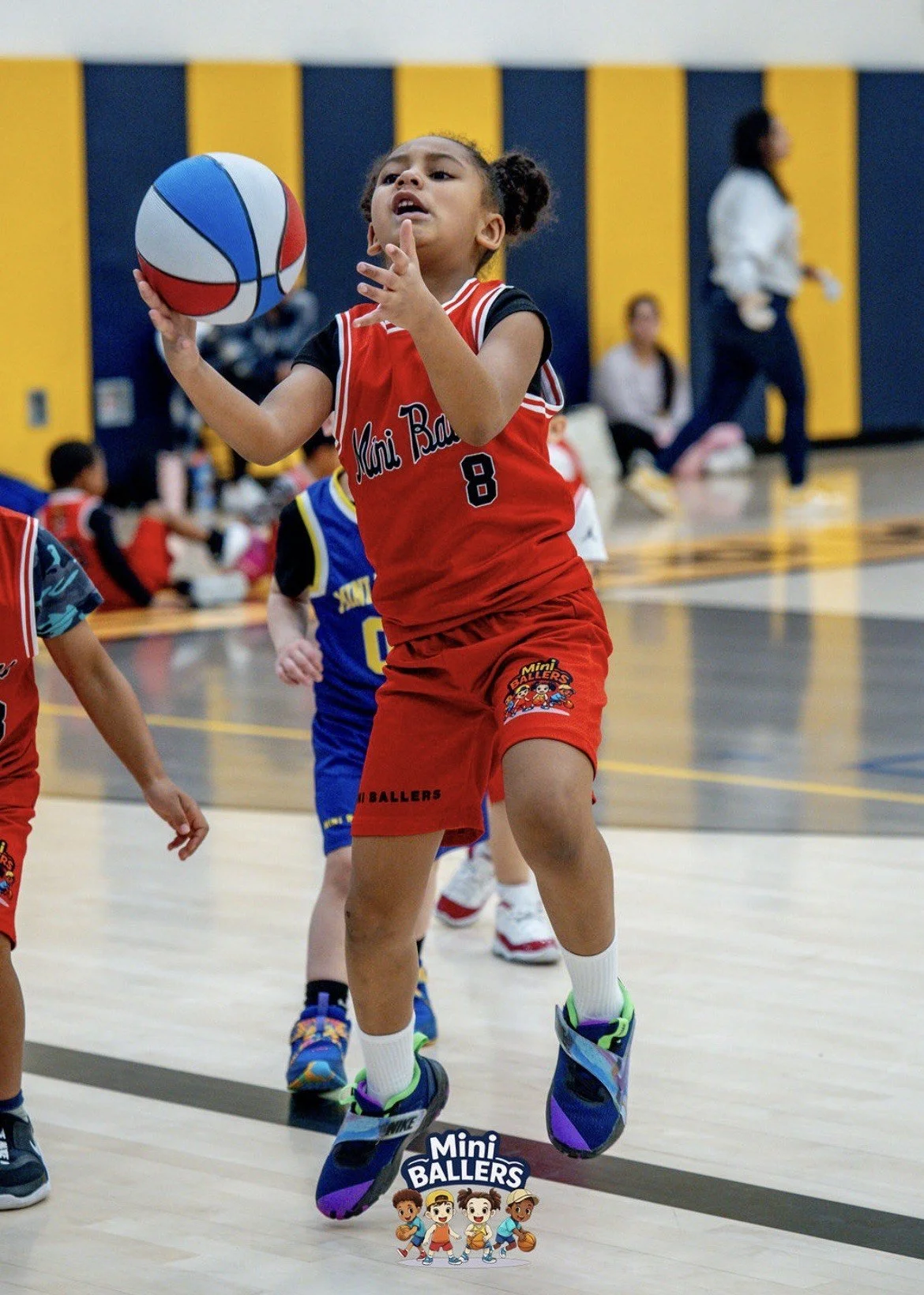 A young girl in a red sports uniform with the number 8 is playing basketball indoors. She is holding a basketball with her right hand, preparing to shoot. Other children are around her, and there are people sitting and walking in the background.
