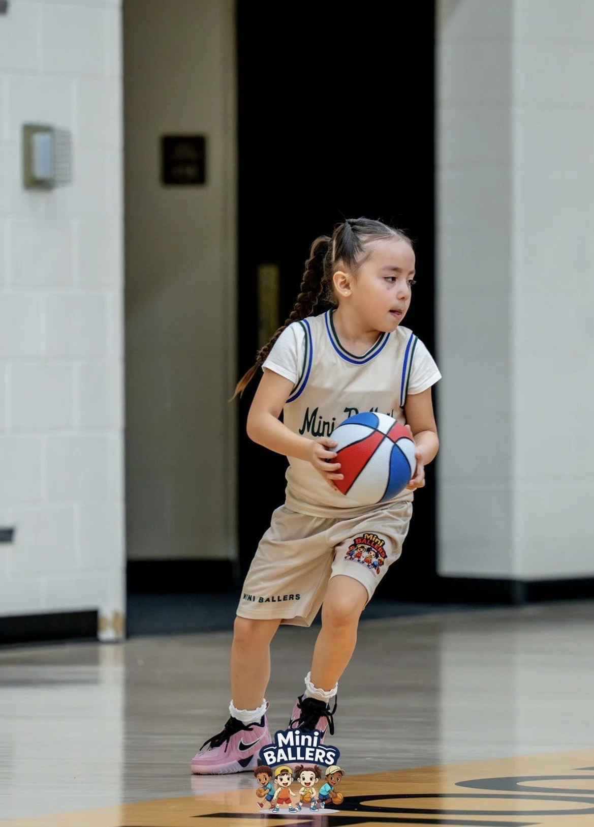 A young girl playing basketball on an indoor court, holding a colorful mini basketball and wearing basketball shorts and sneakers.