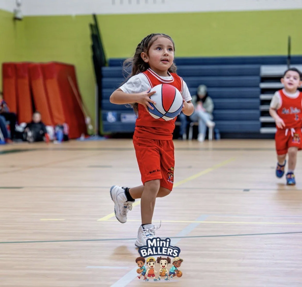 Young girl in a red sports uniform playing basketball indoors, dribbling a basketball, with another girl in a similar uniform in the background.