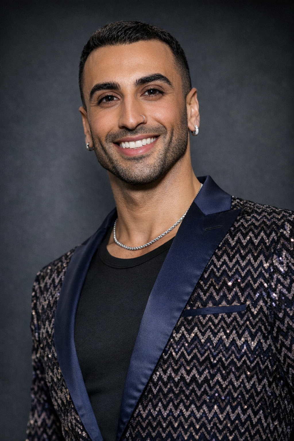 A smiling man with dark hair and a beard, wearing a black shirt, a sequined blazer with a zigzag pattern, jewelry including earrings, a nose ring, and a necklace, posing against a dark background.