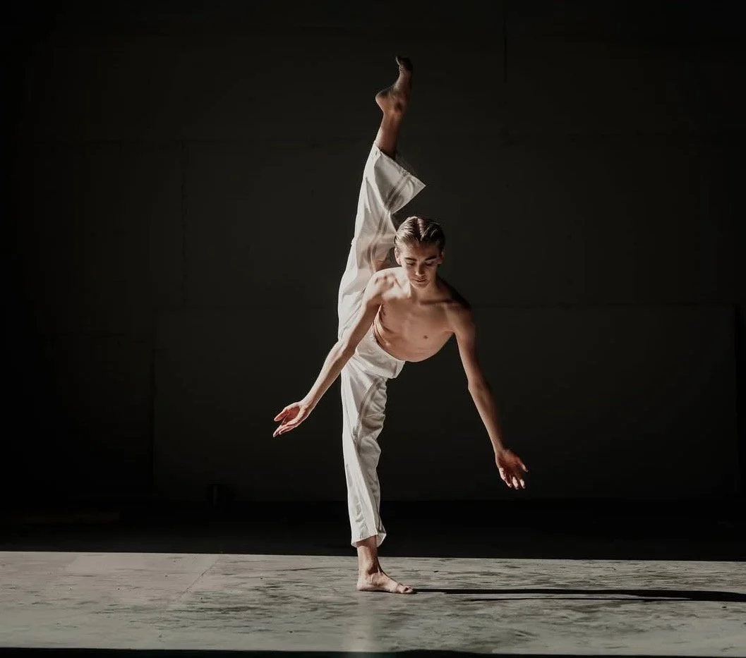 A shirtless male dancer wearing white pants performs a dance move on a dimly lit stage.