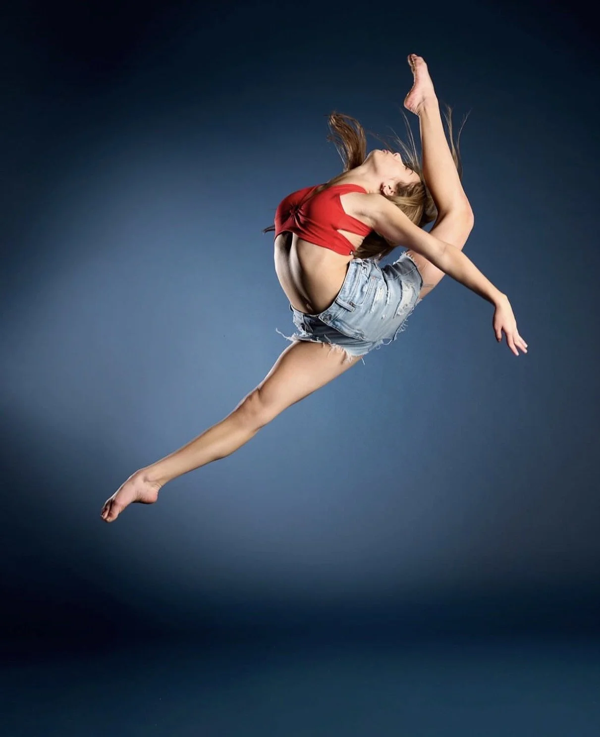 A female dancer in mid-air performing a jump against a dark background, wearing a red crop top and denim shorts.