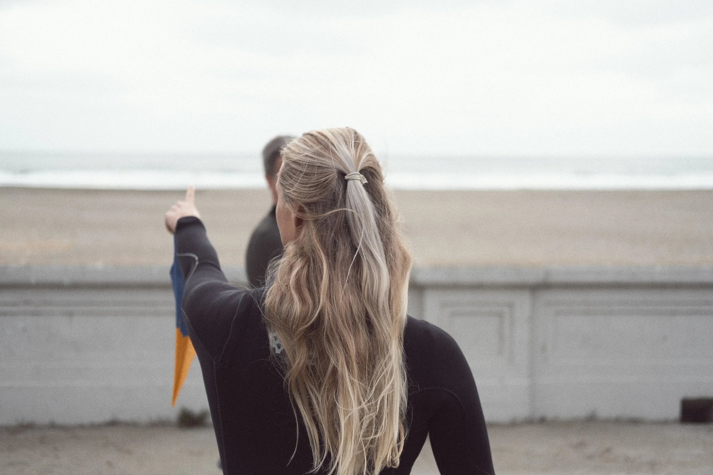 A woman with long blonde hair tied in a ponytail, wearing a black wetsuit, pointing at the ocean with a man in the background at a beach.