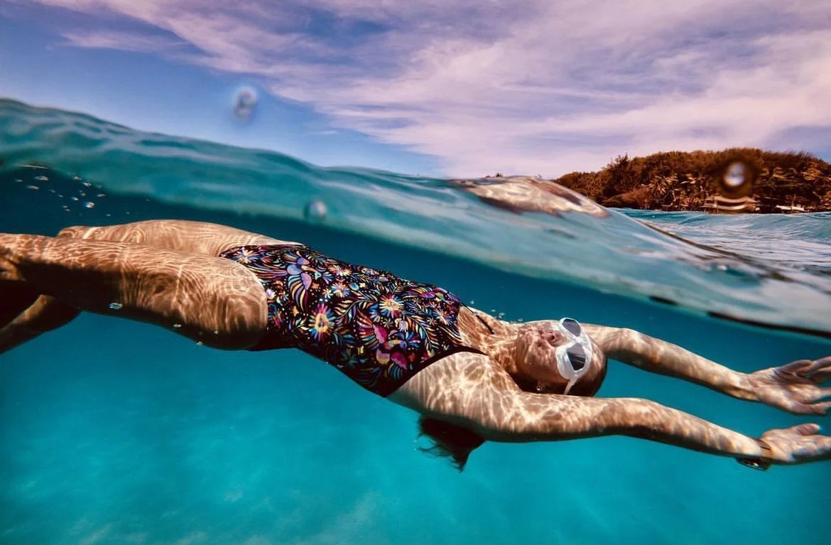 Person swimming in the ocean wearing sunglasses and a colorful swimsuit with a floral pattern, with water and a distant island in the background.