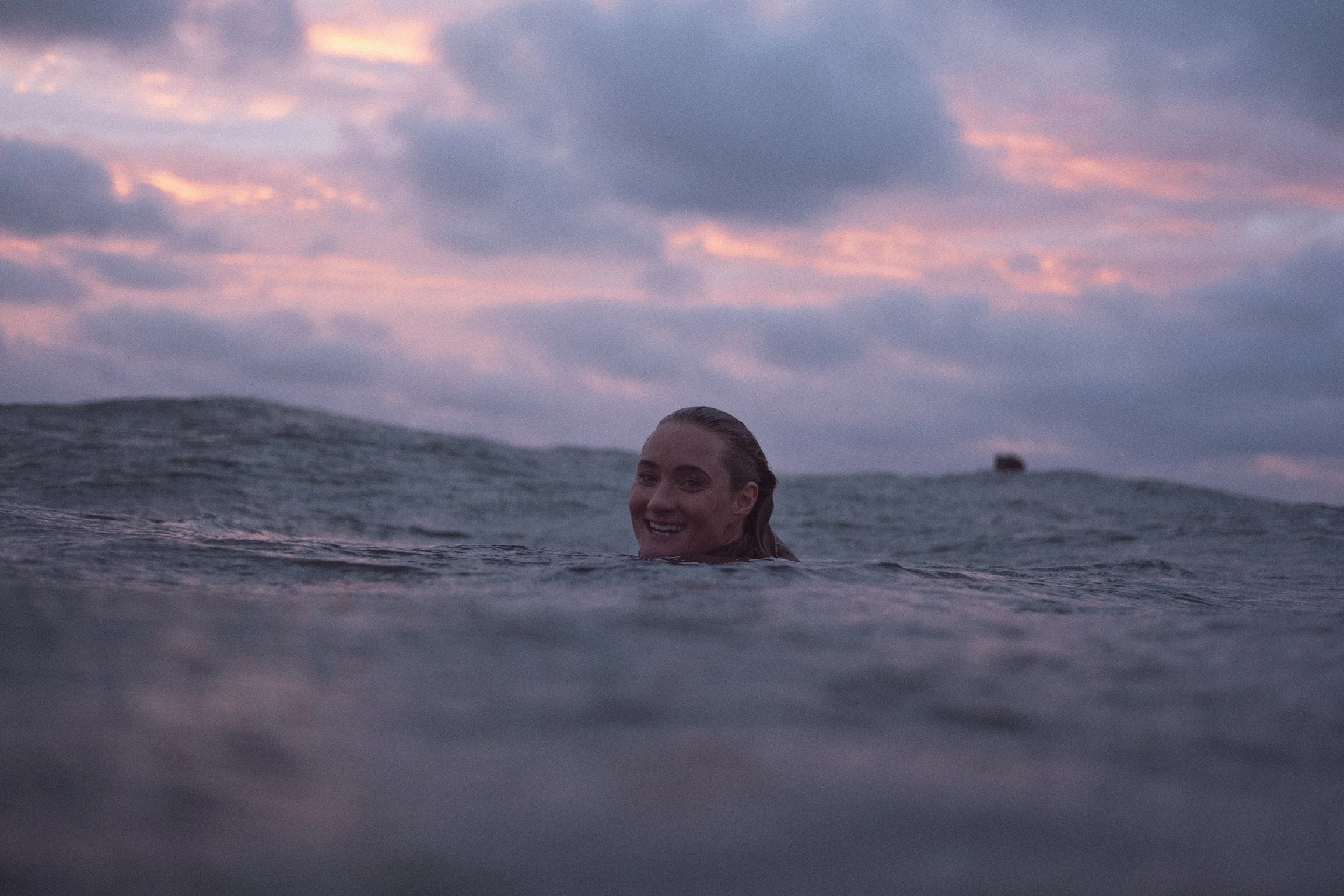 A woman swimming in an ocean with sunset sky and clouds in the background, smiling at the camera.