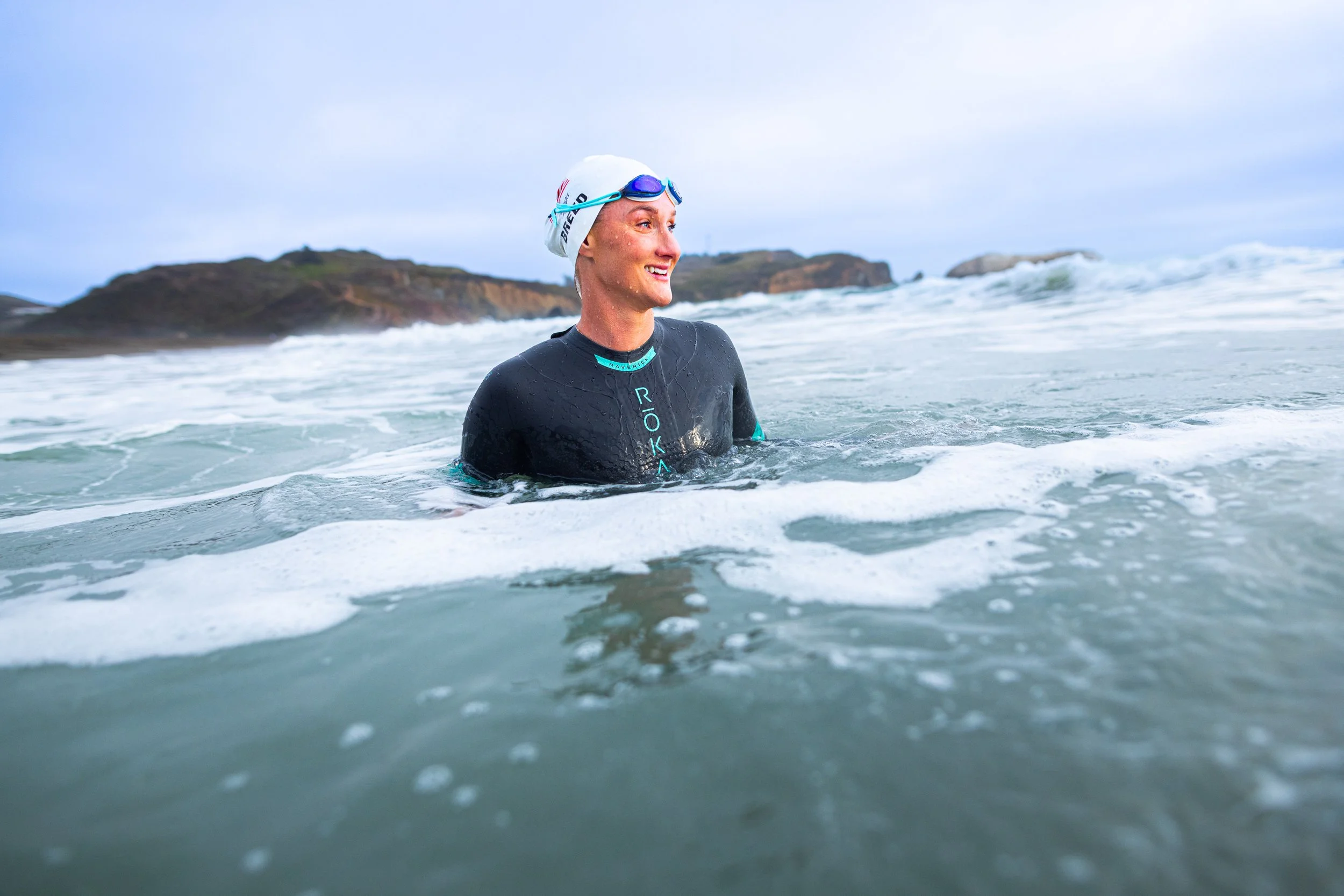 A woman in a wetsuit and swim cap stands in the ocean with waves, smiling, with rocky hills in the background.