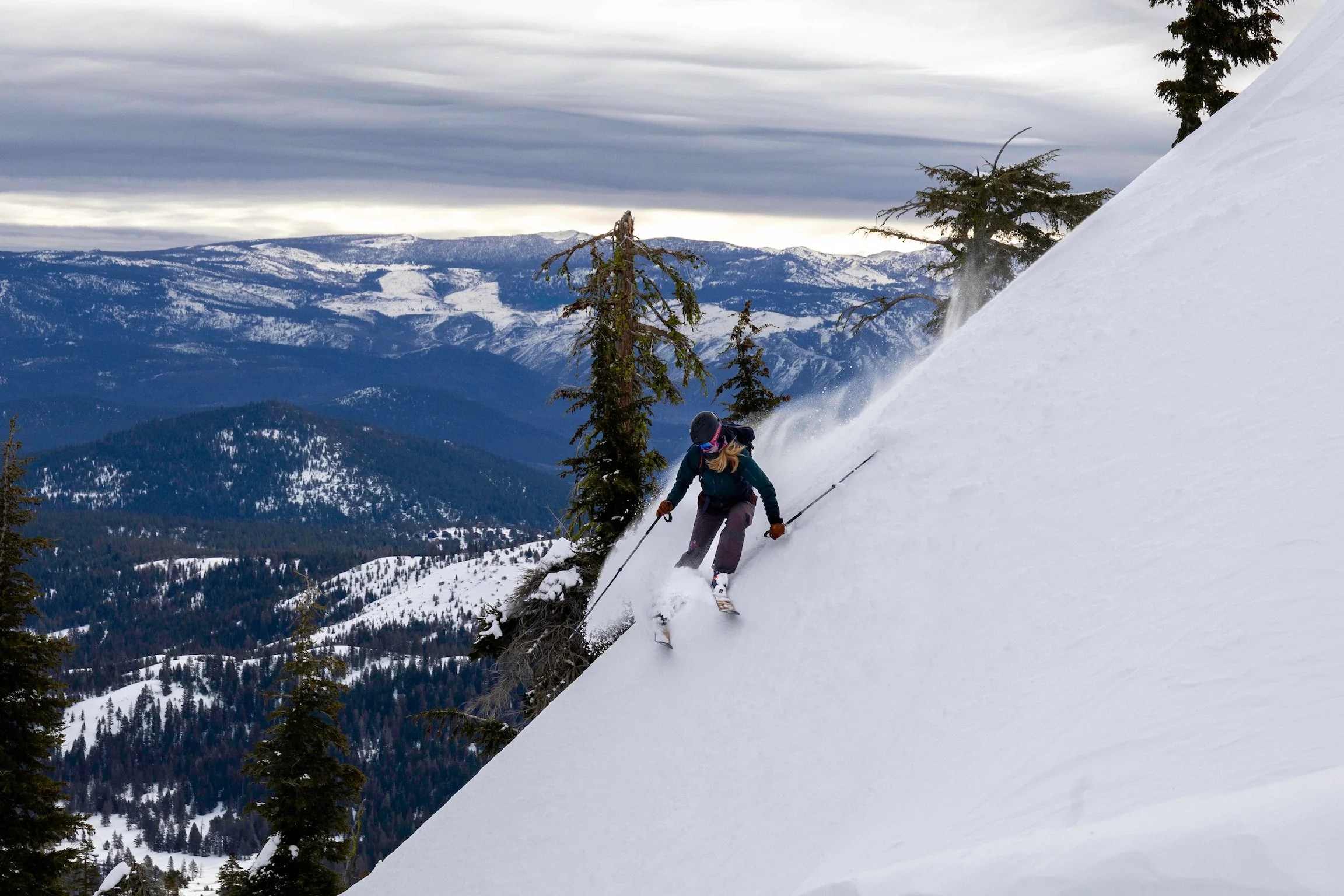 A skier navigating down a snowy mountain slope with trees in the background and mountain ranges under a cloudy sky.