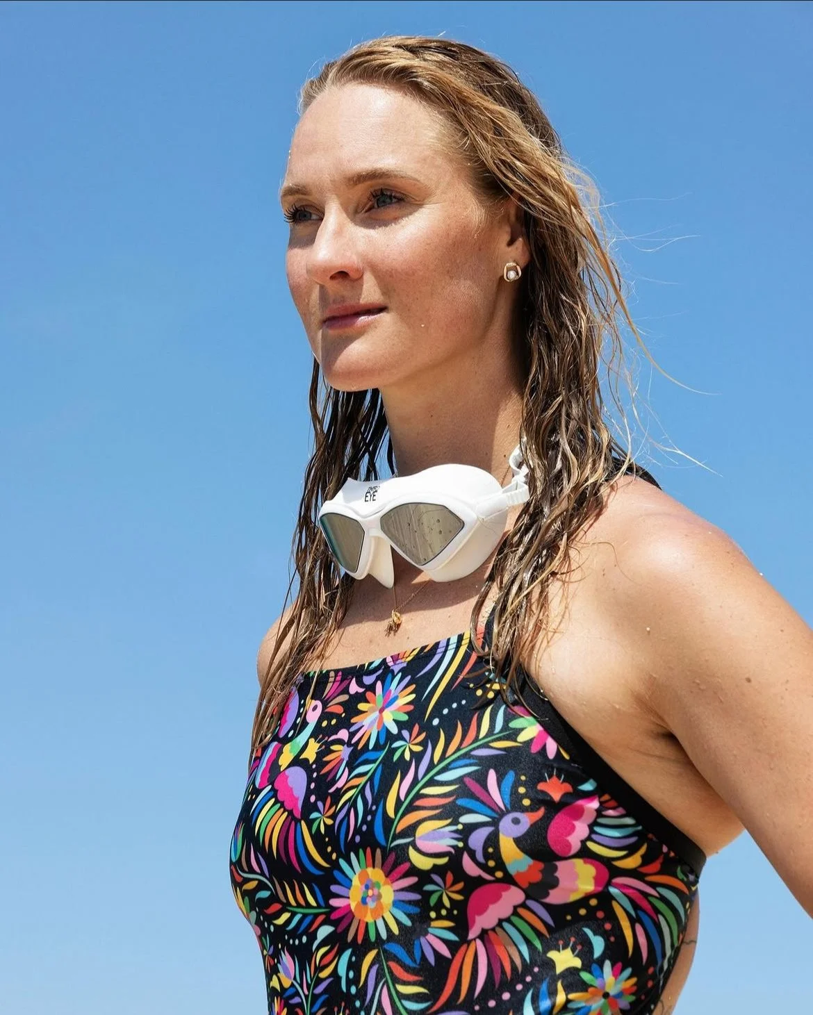 A woman standing outdoors against a clear blue sky, wearing a colorful floral swimsuit, sunglasses around her neck, and jewelry.