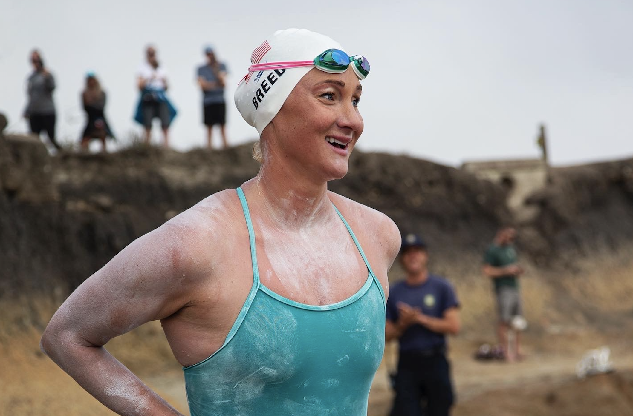 A female swimmer, wearing a white swim cap and teal swimsuit, smiling after a race, with sand or dirt on her skin, in an outdoor setting with spectators in the background.
