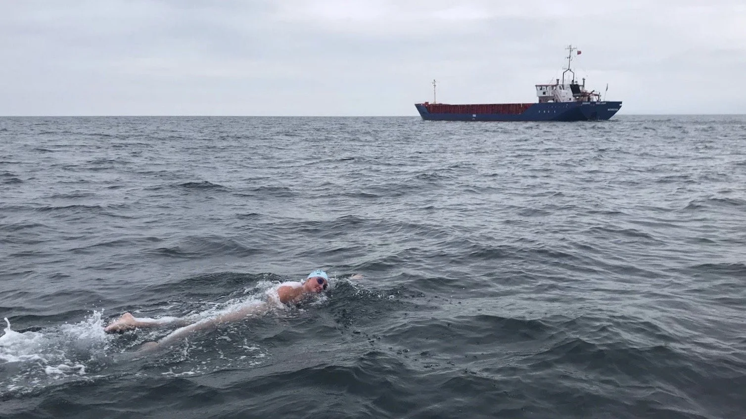 A person swimming in open water near a large cargo ship on a cloudy day.