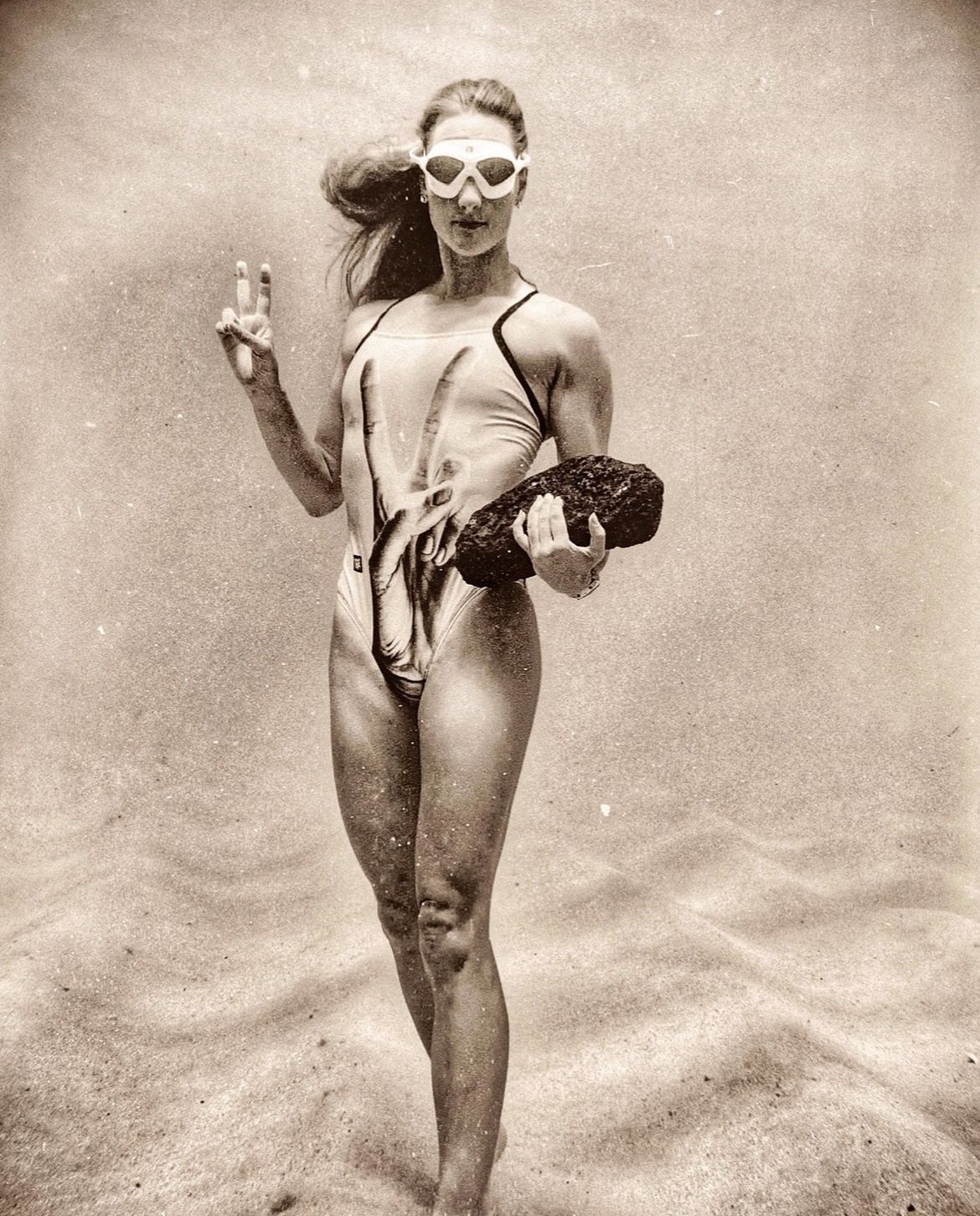 A woman in a swimsuit holding a large rock, wearing goggles, and making a peace sign with her right hand, standing on sand.