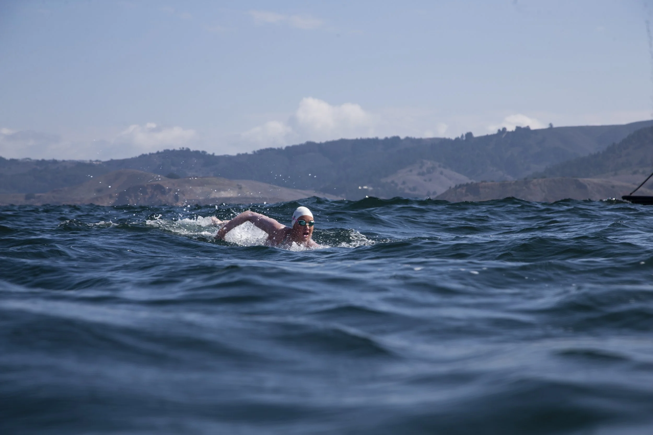 Swimmer in a white swim cap and goggles swimming in open water, with hills and sky in the background.
