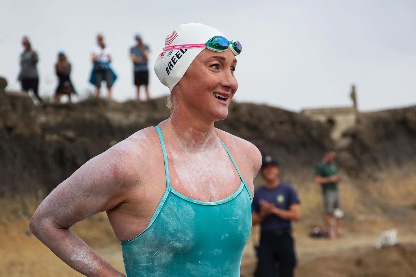 A smiling female swimmer in a turquoise swimsuit and white swimming cap with goggles resting on her forehead, standing at an outdoor swimming event with a dirt or rock hill and several spectators in the background.