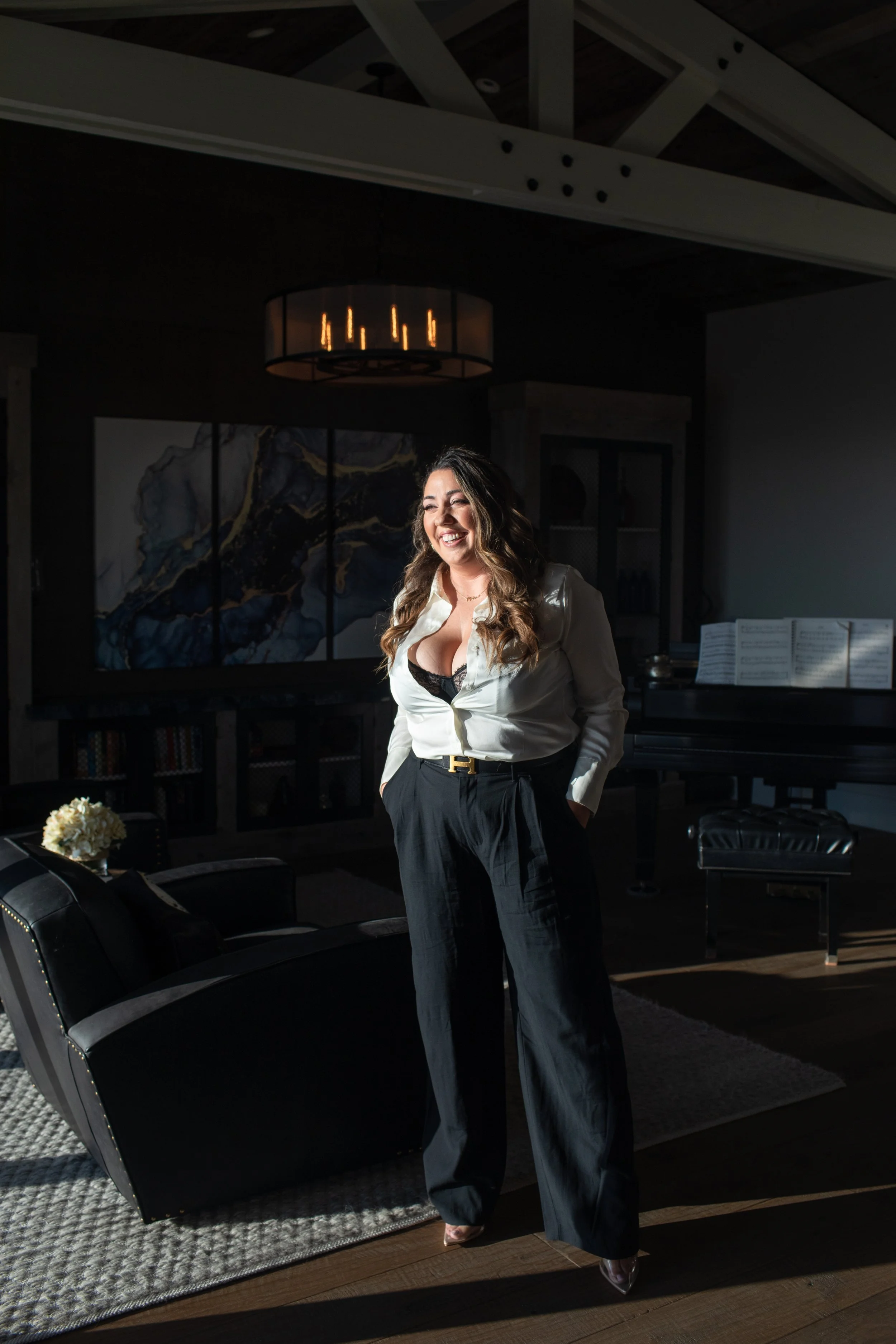 A woman standing in a dimly lit living room, smiling, wearing a white blouse, black dress pants, and heels, with sunlight streaming in.