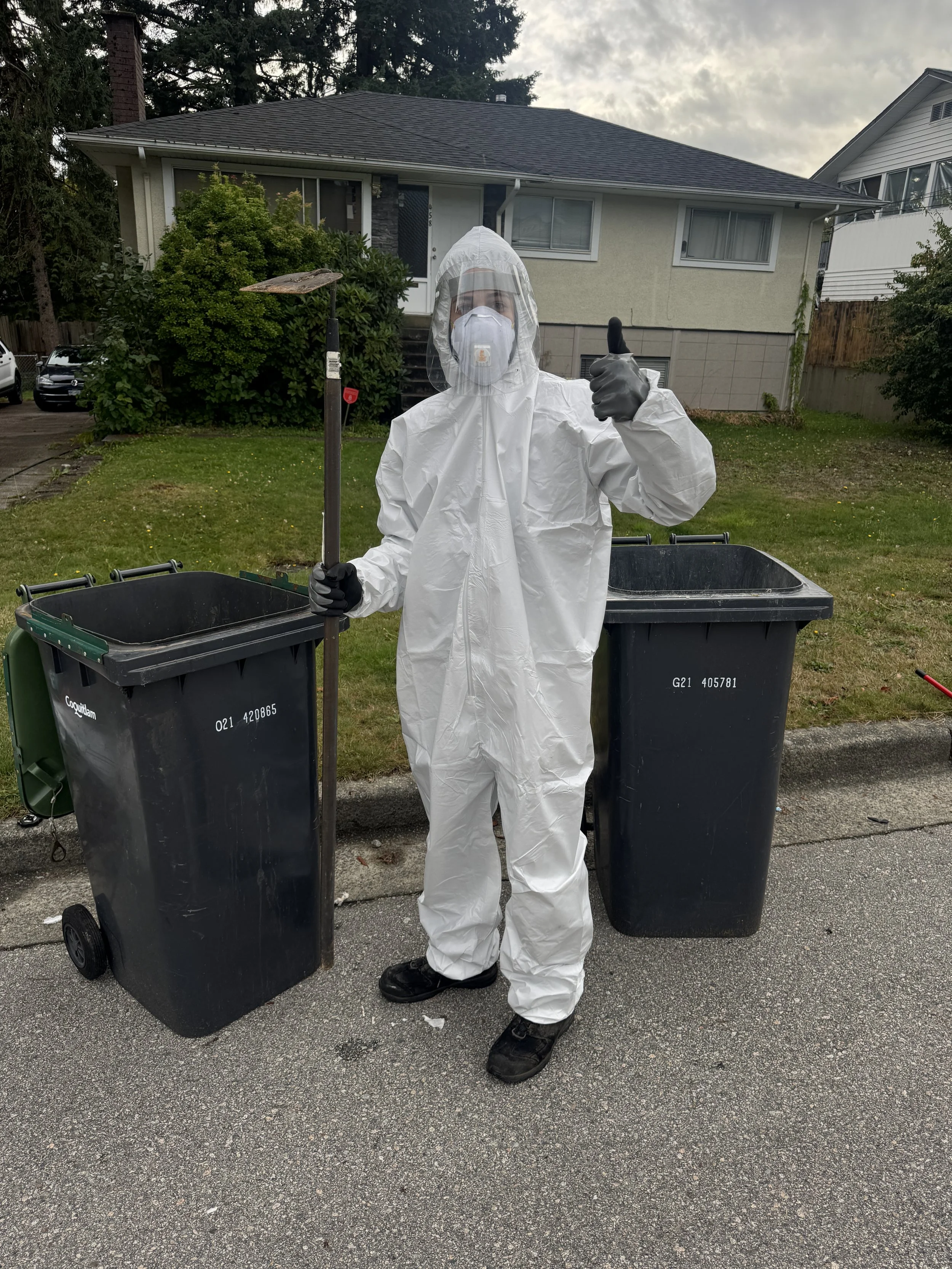 Person wearing full protective gear including a white hazmat suit, gloves, mask, and face shield, standing outdoors between two black trash bins, holding a trash picker and giving a thumbs-up gesture.