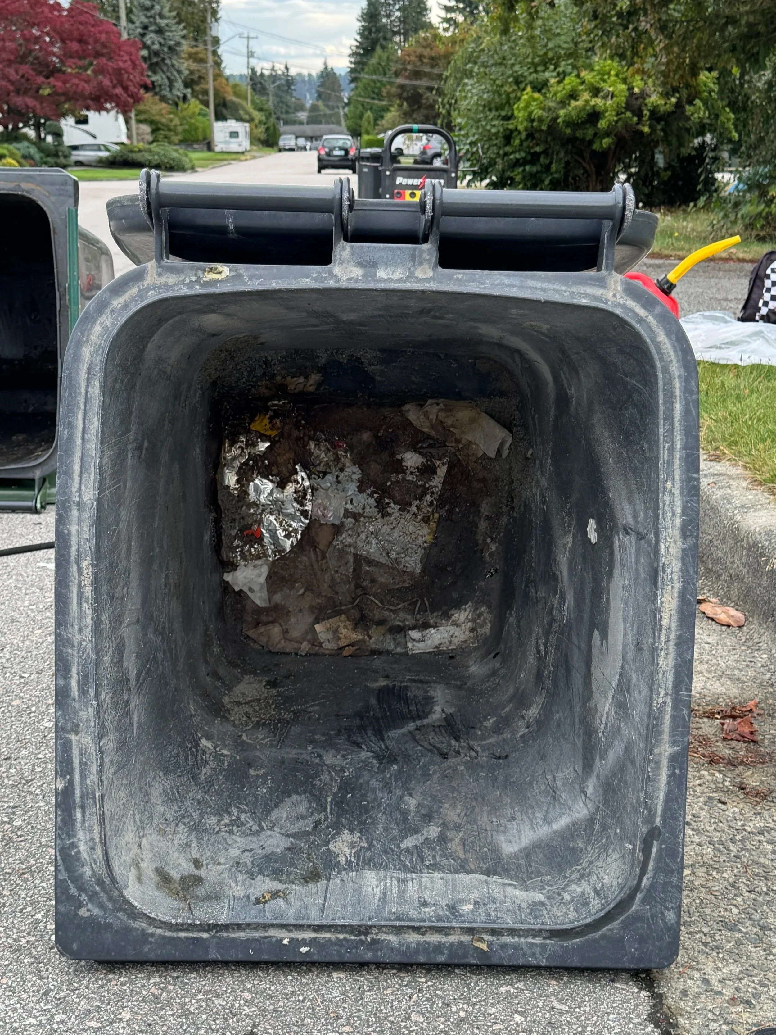 Empty black trash bin with some trash and debris inside, outside on the pavement in a residential area.