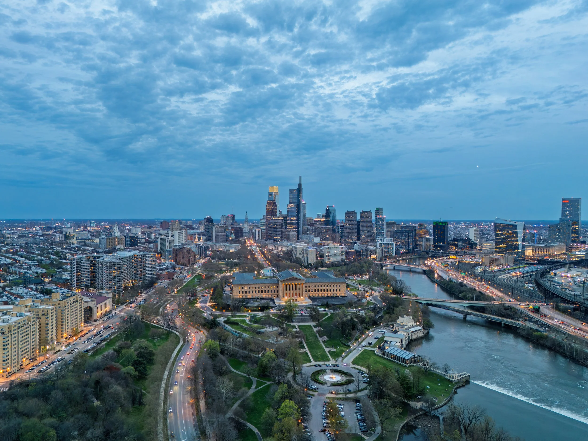 Aerial view of a city skyline at dusk with high-rise buildings, a river, and a park with walkways.