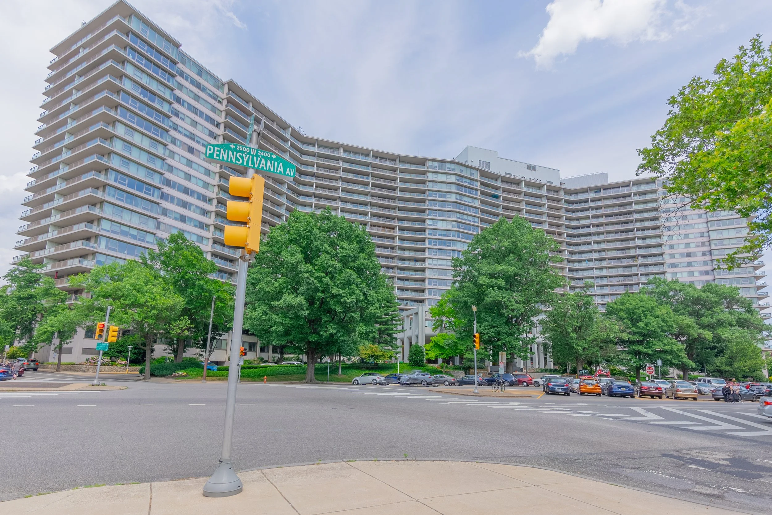 A large residential building with numerous balconies, featuring a curved design, on a city street corner. There are green trees, parked cars, and traffic lights. Street signs indicate the intersection of Pennsylvania Avenue and 24th Street.