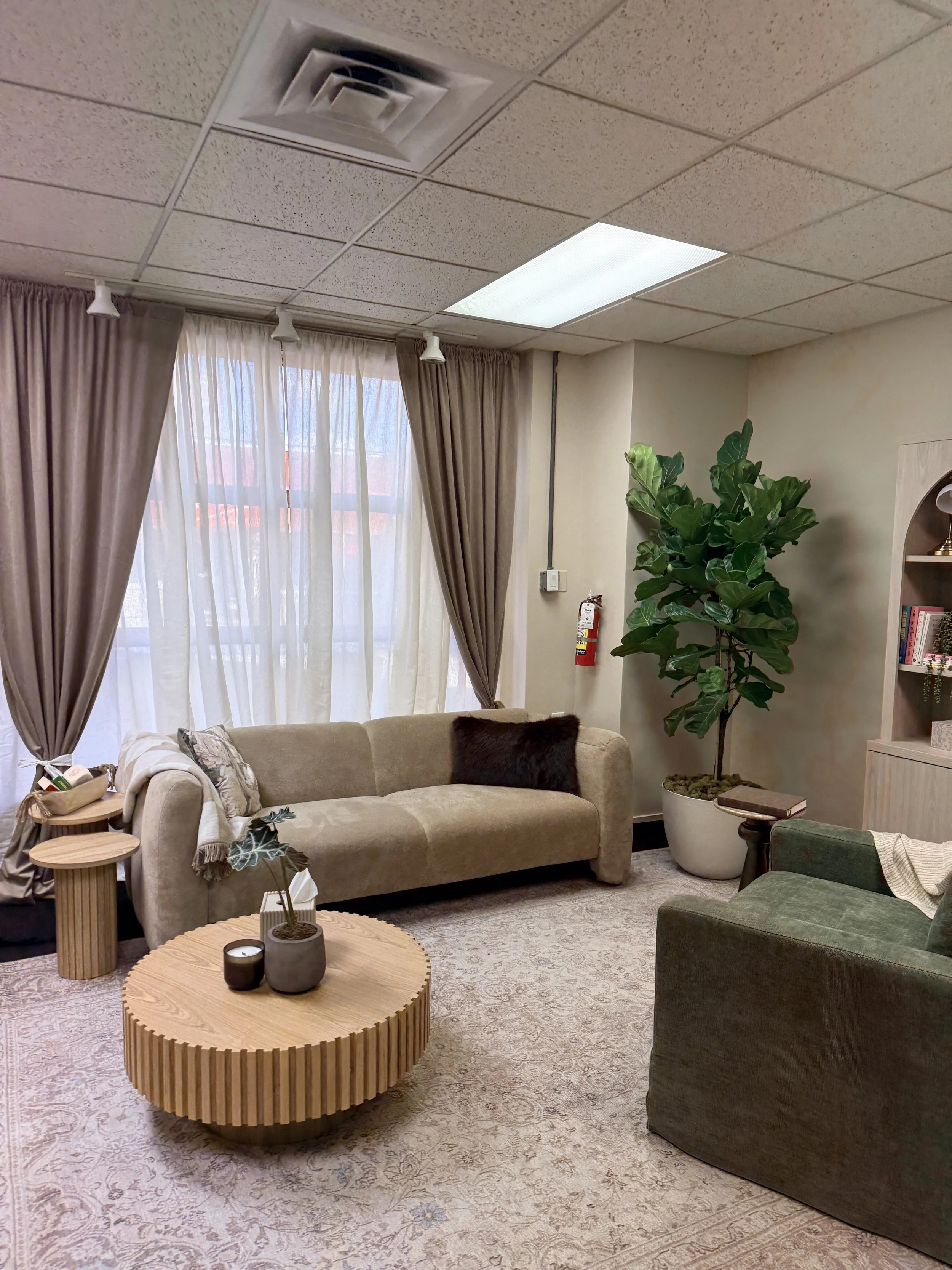 A room with a beige couch, a green armchair, a wooden coffee table with candles, a large potted plant, a window with beige curtains, and a ceiling with fluorescent lights and an air vent.