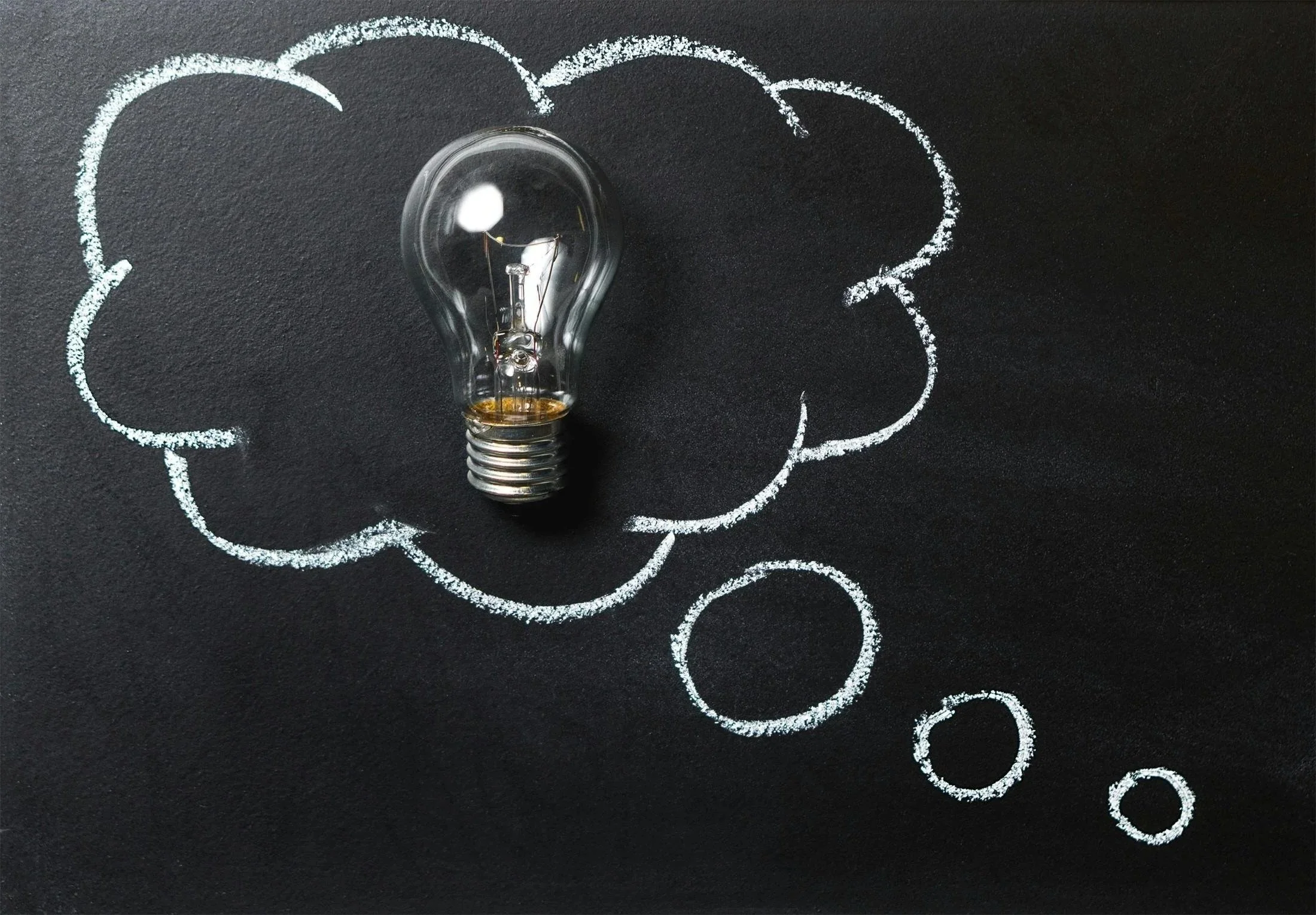 A light bulb placed on a black surface with a hand-drawn thought bubble around it, created with white chalk.