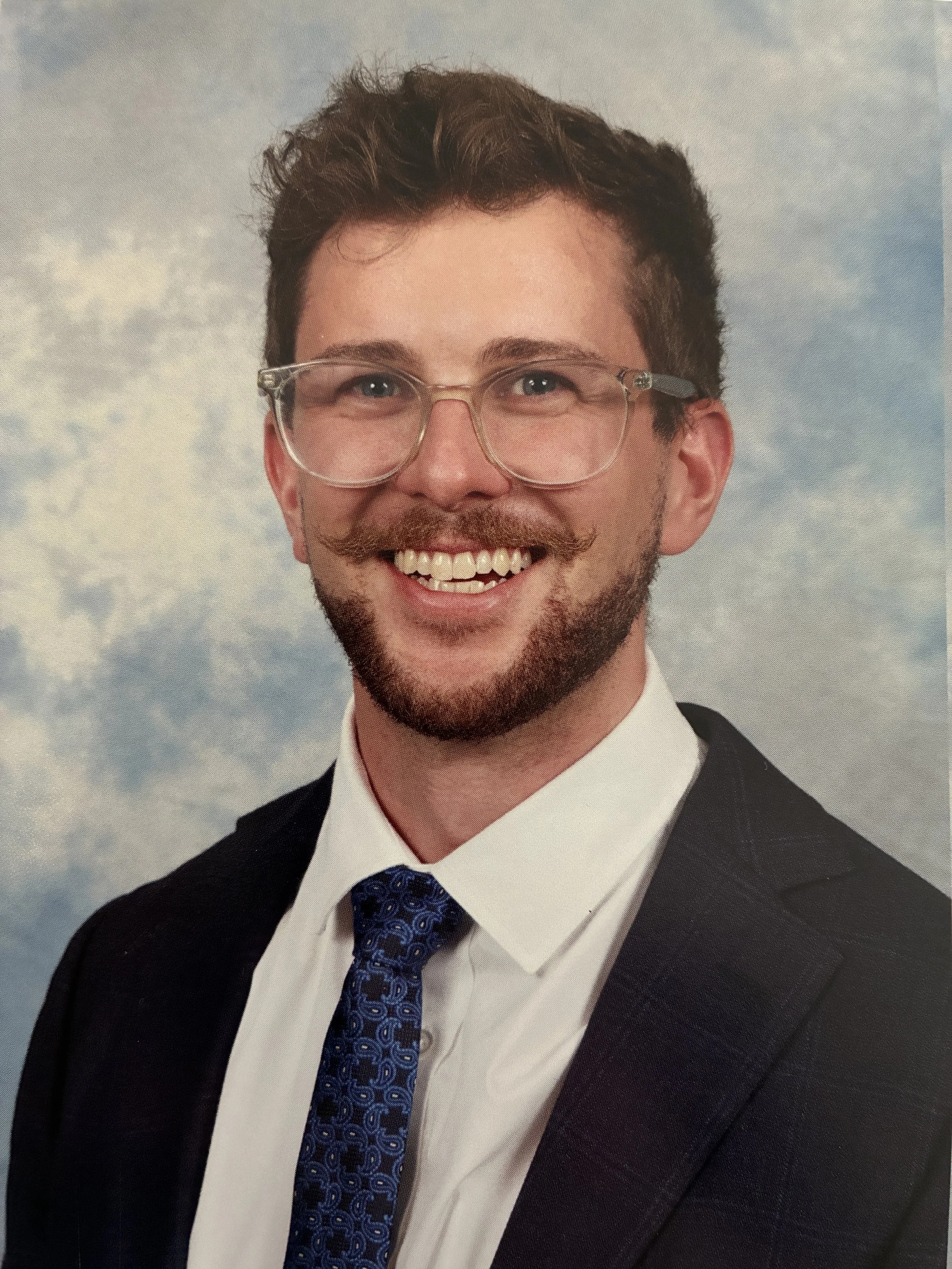 Portrait of a smiling man with glasses, a beard, and a mustache, wearing a suit, white shirt, and a patterned blue tie, against a cloudy sky background.