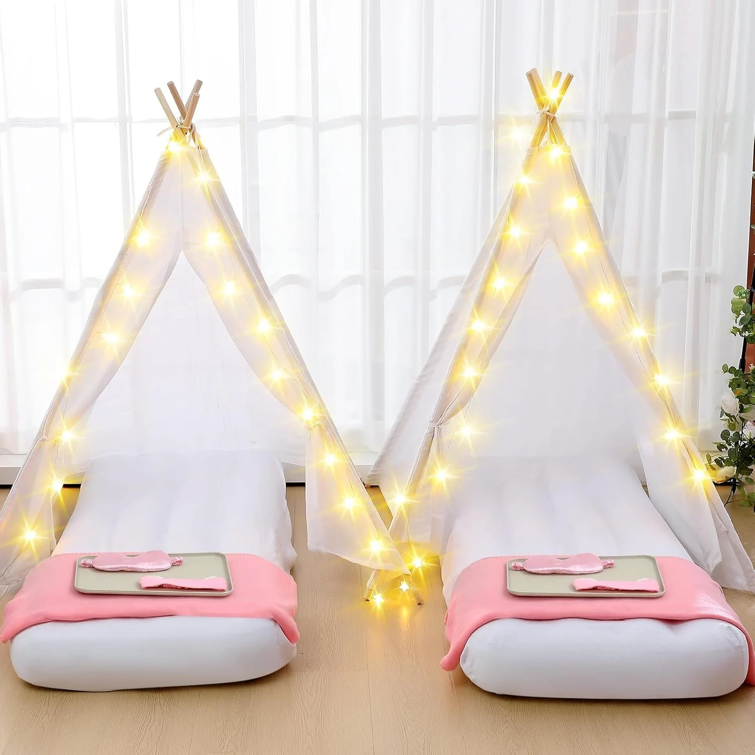 Two indoor beds designed as cozy teepees with string lights, pink blankets, and tray tables, positioned on a wooden floor in front of large windows with sheer curtains and a plant on the side.
