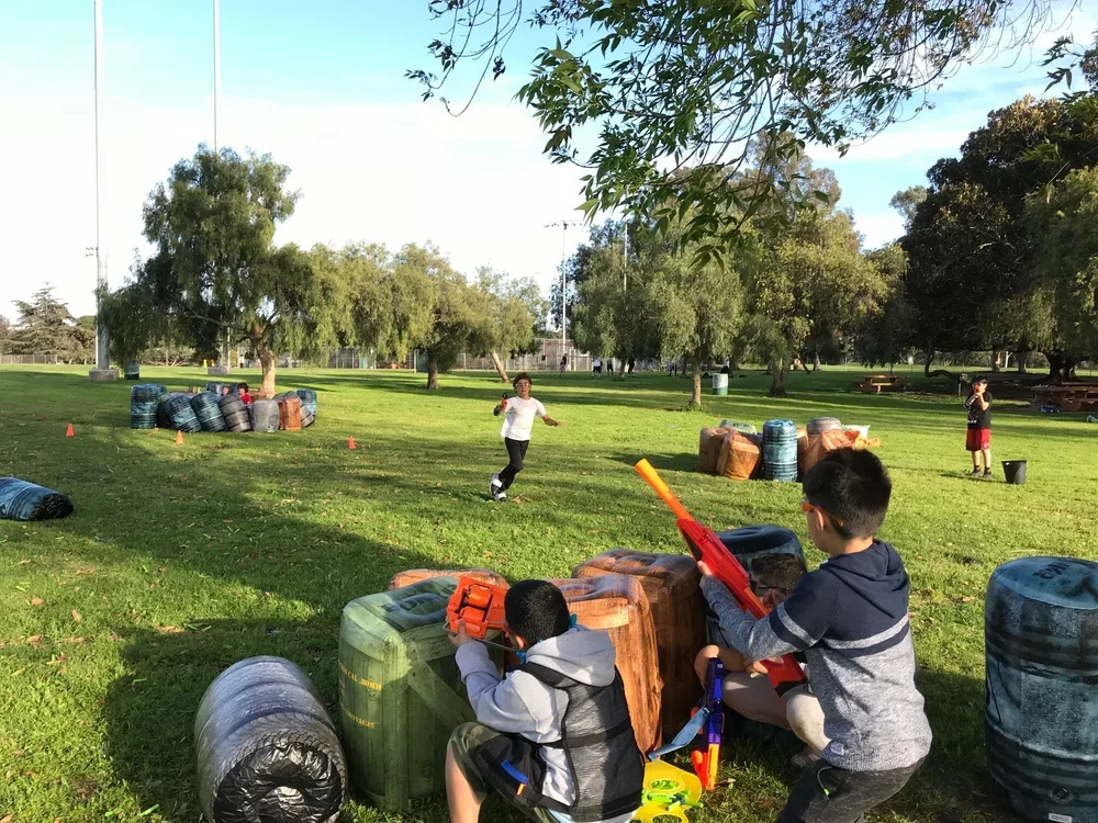 Children playing in a park with toy Nerf guns, hiding behind large inflatable barriers made to look like barrels.