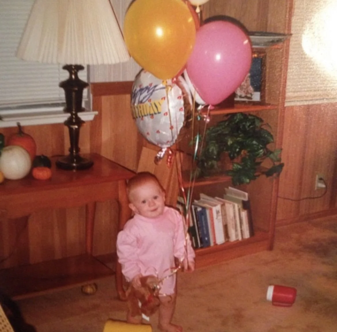 A young child holding a bunch of colorful balloons in a living room with wooden furniture and bookshelves.