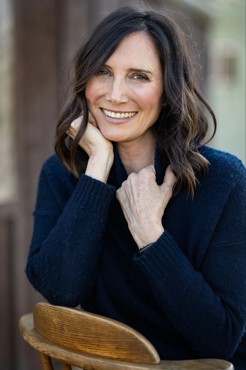 A woman with shoulder-length dark brown hair smiling at the camera, resting her chin on her hand, wearing a navy blue sweater, sitting in a wooden chair outdoors.