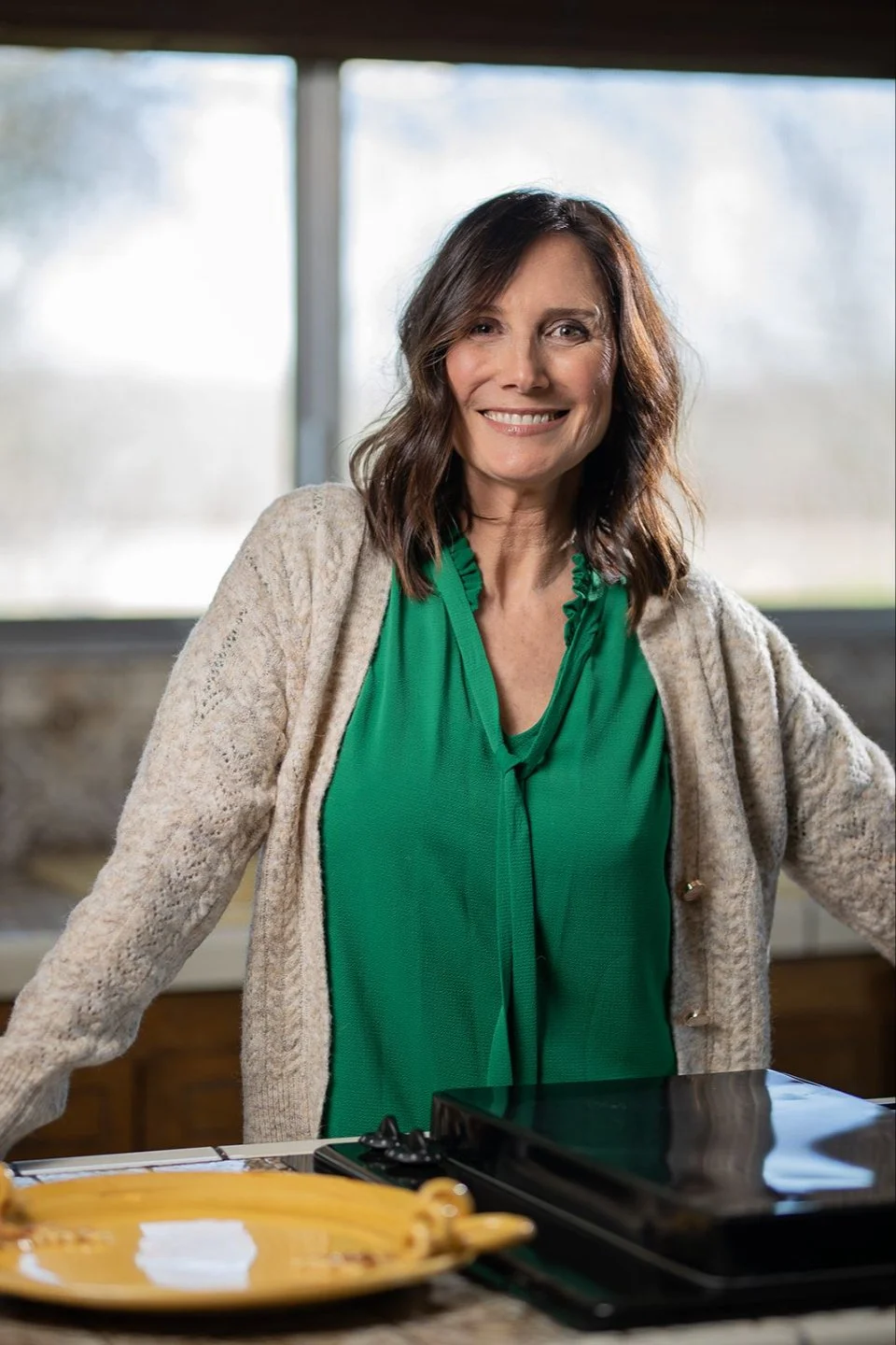 A smiling woman with shoulder-length brown hair, wearing a green blouse and a beige cardigan, standing in a kitchen with a large window in the background.