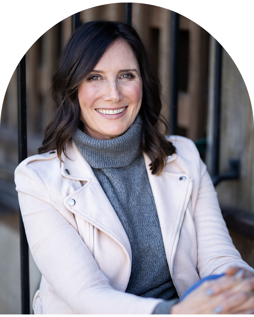 A woman with shoulder-length dark hair, smiling, wearing a gray turtleneck sweater under a light-colored jacket, posing outdoors.