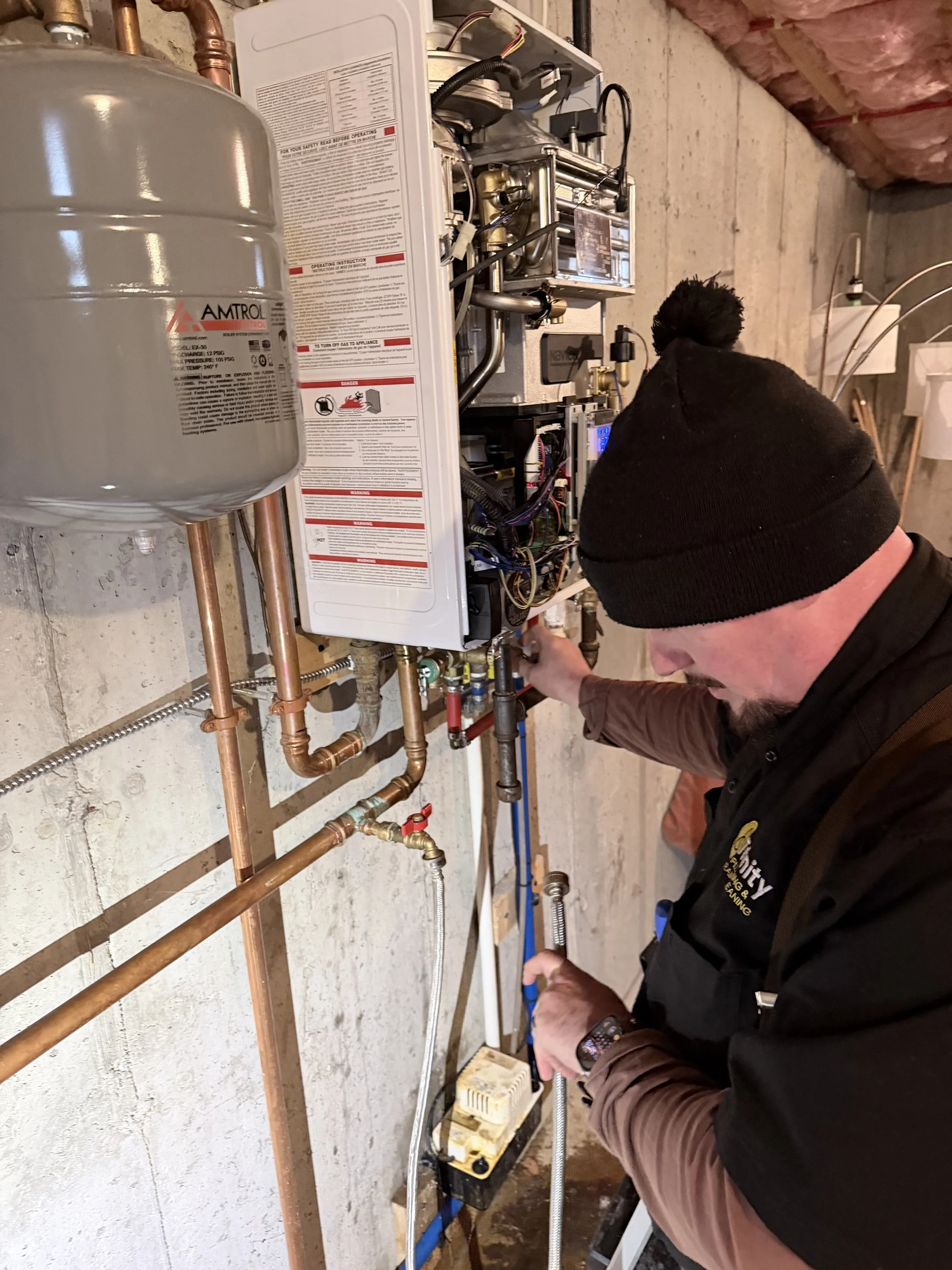 A technician working on a water heater or boiler system, connecting or adjusting pipes in a basement with exposed concrete walls and ceiling insulation.